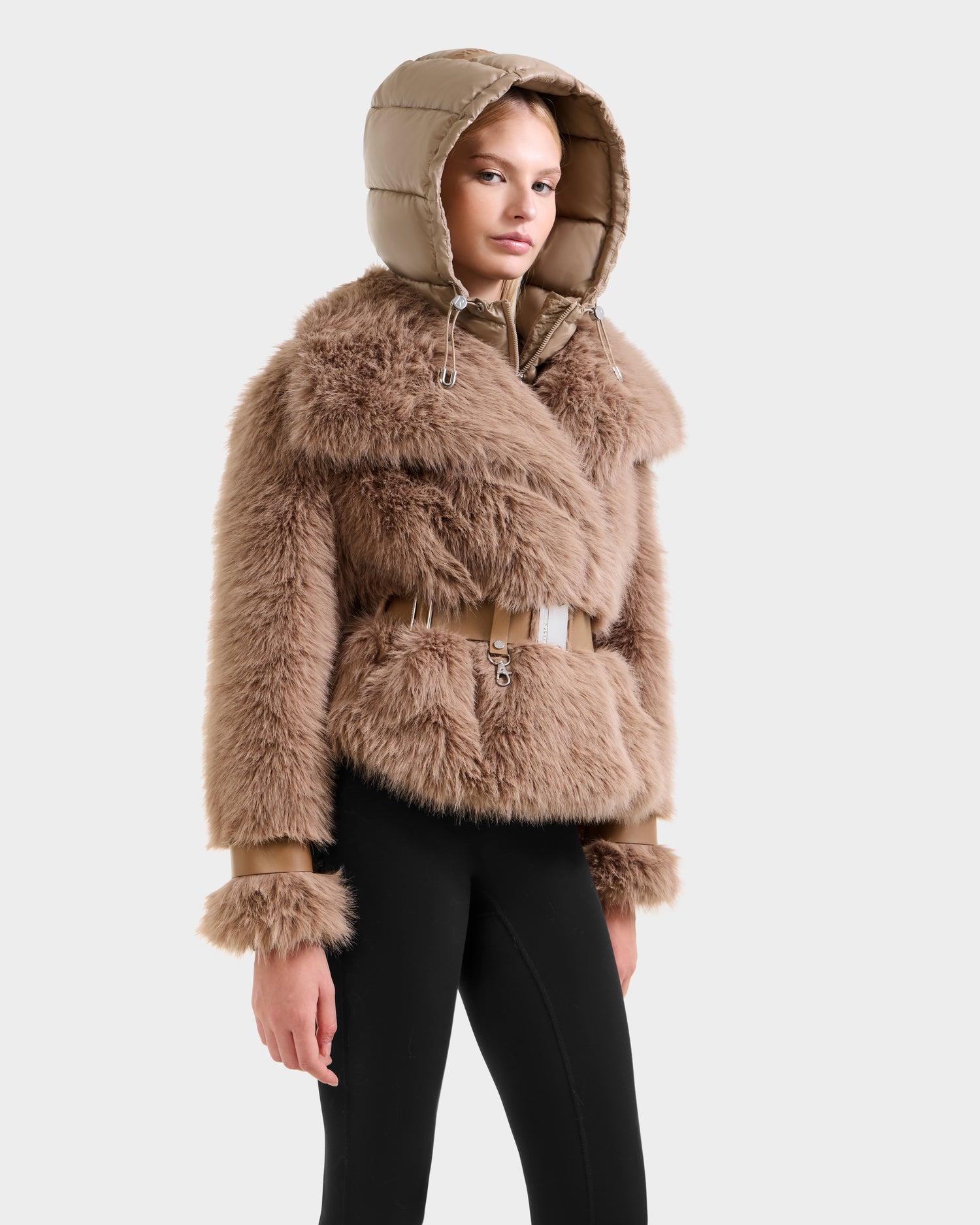 A person models the Rudsak CHLOE - 8125619 Camel women's faux fur jacket with belted waist and hood, paired with black leggings against a light gray background—an effortlessly chic winter look.