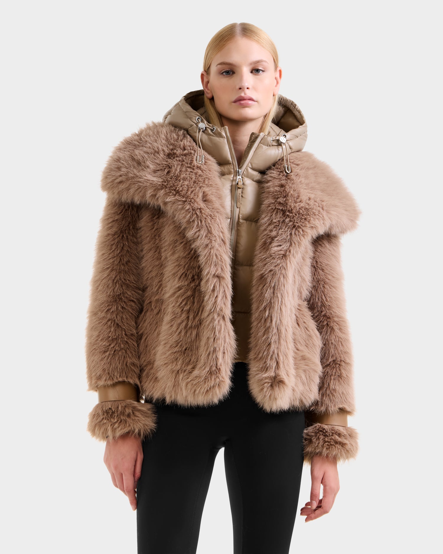 A woman in the Rudsak CHLOE - 8125619 Camel faux fur jacket over a beige hooded puffer, with black pants, stands against a plain light gray background—an effortlessly chic winter look.