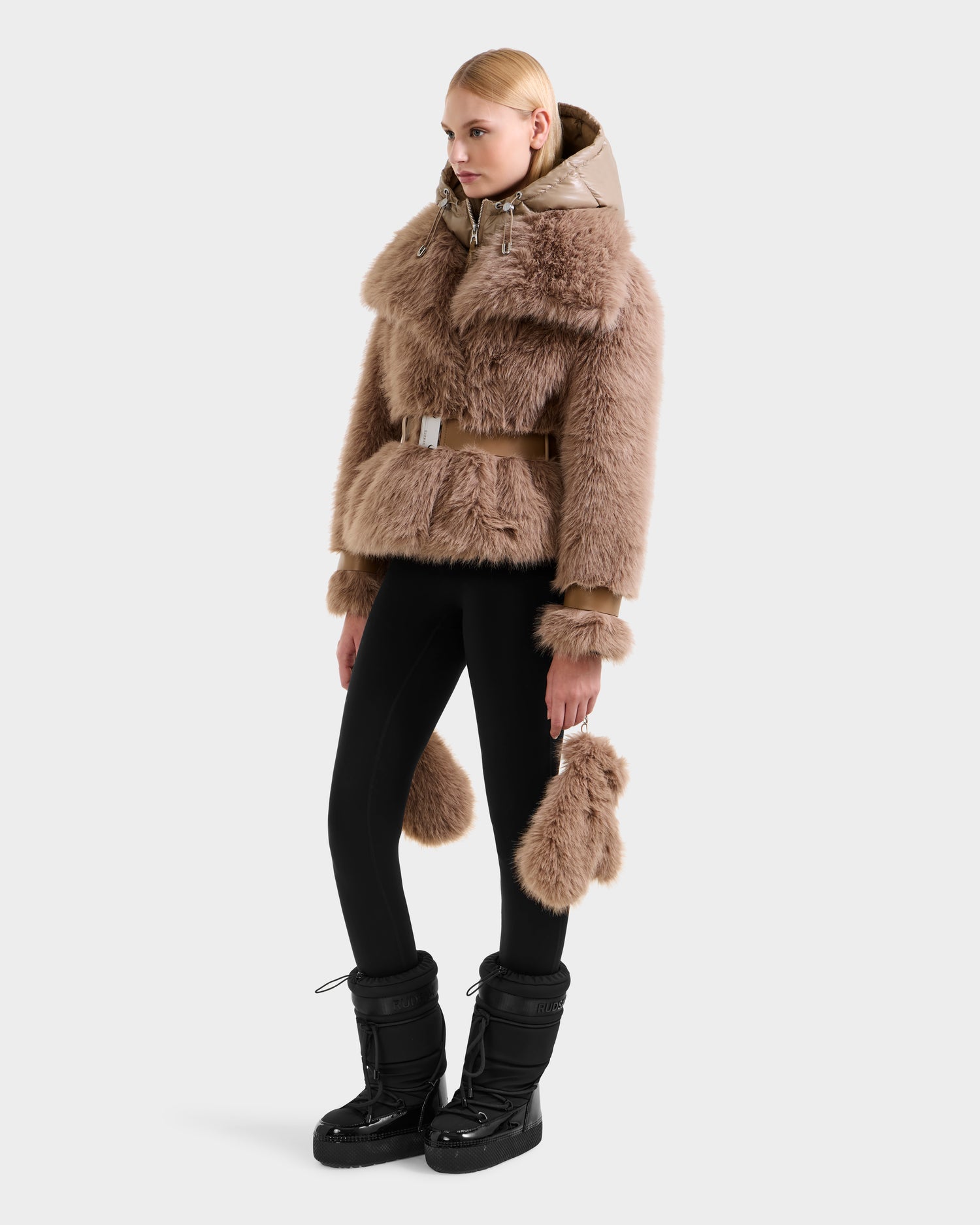 A woman models the CHLOE - 8125619 Camel faux fur jacket by Rudsak, featuring a hood and belt. She pairs it with black leggings, black snow boots, and a matching accessory for a chic winter look.