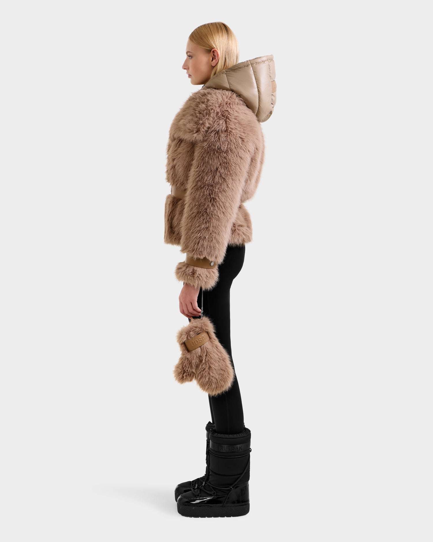 A person in profile models the Rudsak CHLOE - 8125619 Camel women's faux fur jacket with a shiny hood, black leggings, winter boots, and a matching accessory, highlighting chic winter fashion against a plain light background.