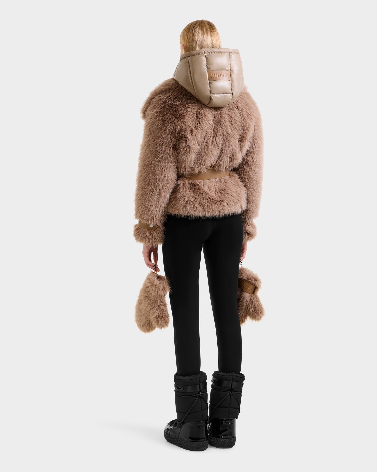 A person with blonde hair, seen from behind, models chic winter style in the Rudsak CHLOE - 8125619 Camel women’s faux fur jacket with a shiny hood, paired with black pants, black winter boots, and a matching tan faux fur accessory.