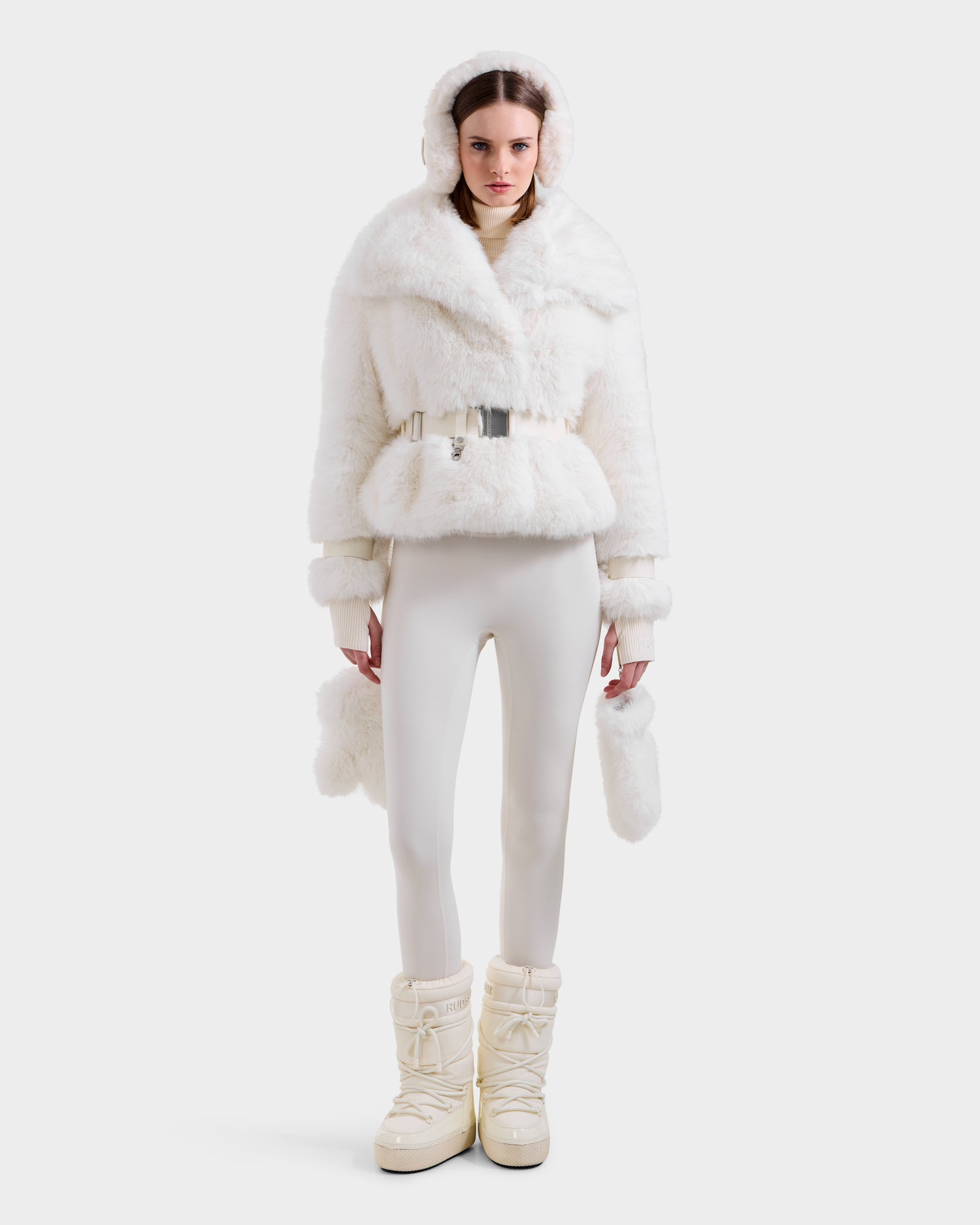 A woman stands against a white background, showcasing winter fashion in Rudsak's CHLOE - 8125619 Iceland faux fur jacket with hood and belt, paired with fitted pants, fluffy mittens, and chunky snow boots.