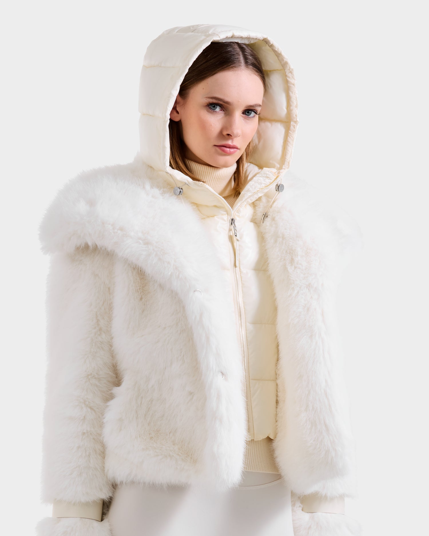 A woman models winter style in the Rudsak CHLOE - 8125619 Iceland faux fur jacket, layered over a cream quilted hooded vest and light turtleneck, set against a plain light background.