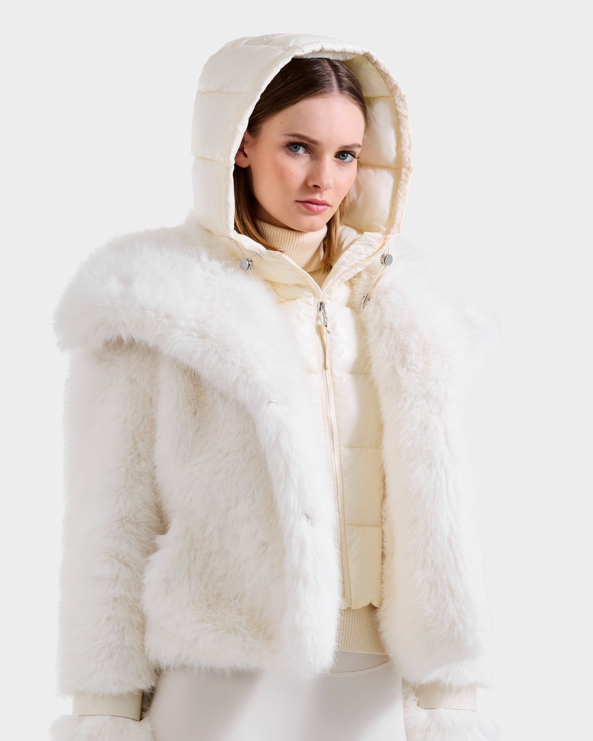 A woman models winter style in the Rudsak CHLOE - 8125619 Iceland faux fur jacket, layered over a cream quilted hooded vest and light turtleneck, set against a plain light background.