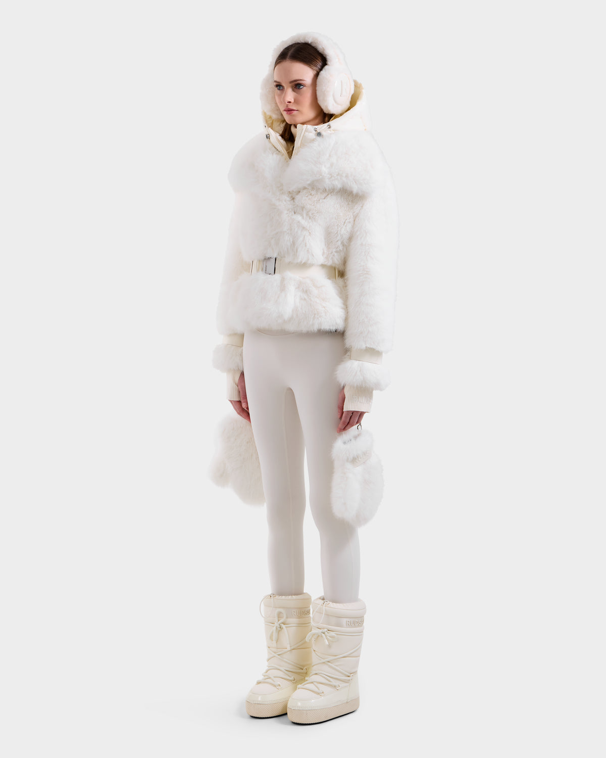 A woman stands against a white background wearing the Rudsak CHLOE - 8125619 Iceland women's faux fur jacket with a fluffy hood, paired with white earmuffs, gloves, cream leggings, and off-white snow boots. She has a neutral expression.