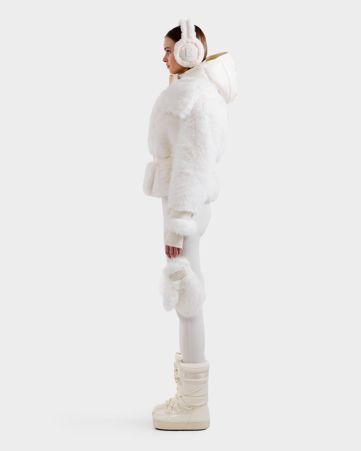 A woman in profile models the Rudsak CHLOE - 8125619 Iceland faux fur hooded jacket with matching earmuffs, white pants, and off-white boots, accessorized with a white faux fur piece—perfect winter style against a plain background.