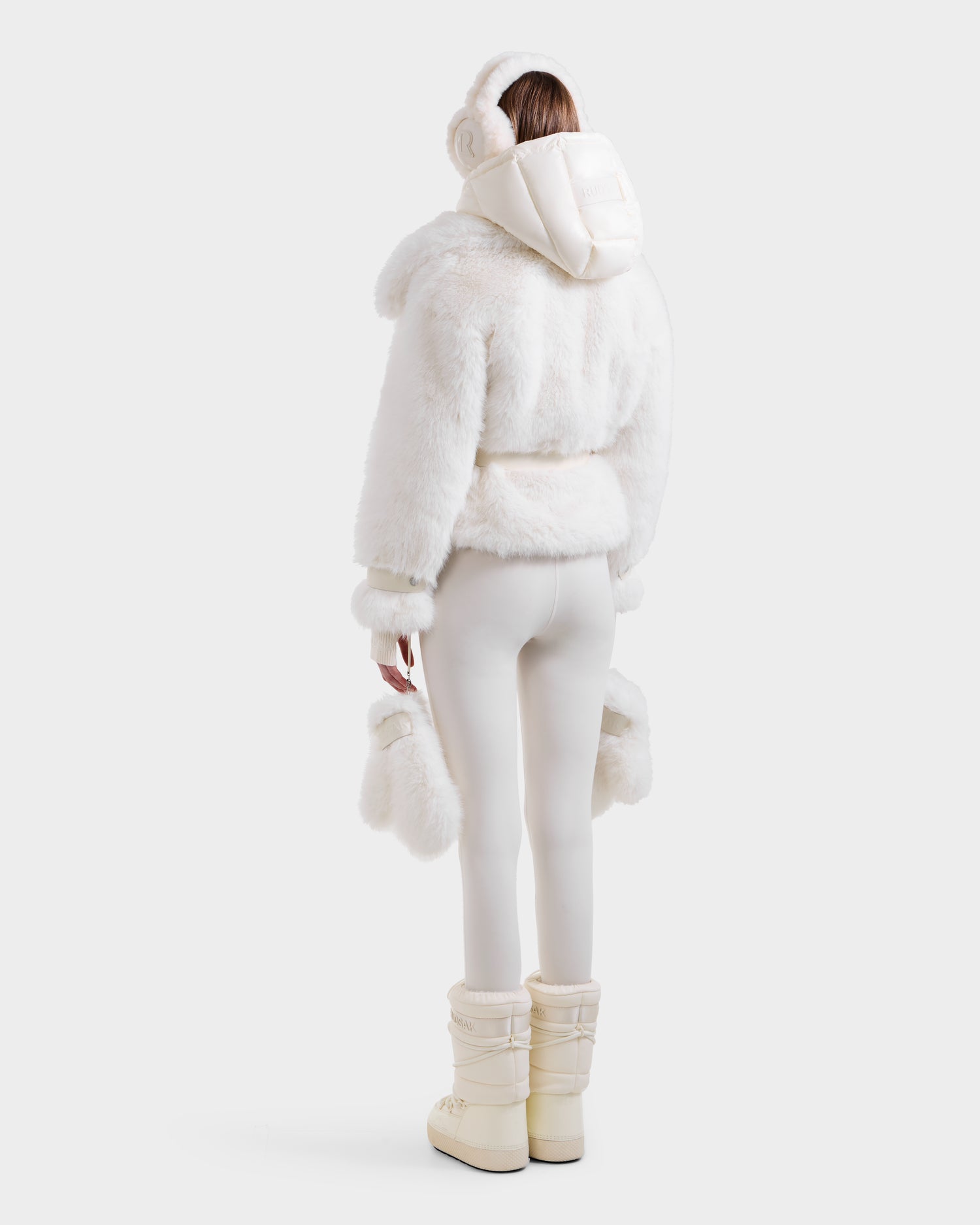 A person faces away, modeling Rudsak's CHLOE - 8125619 Iceland faux fur jacket with hood, white earmuffs, fitted leggings, off-white boots, and matching mittens in a minimalist studio.