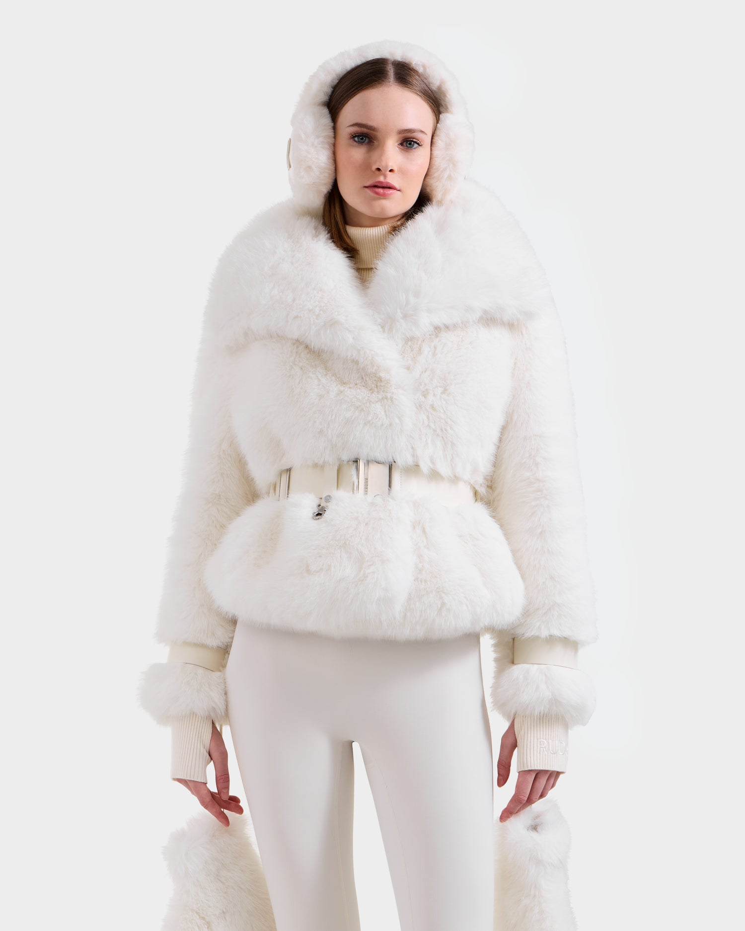 A woman models the CHLOE - 8125619 Iceland faux fur hooded jacket by Rudsak, paired with matching earmuffs and fitted white pants, creating a chic winter look against a plain white background.