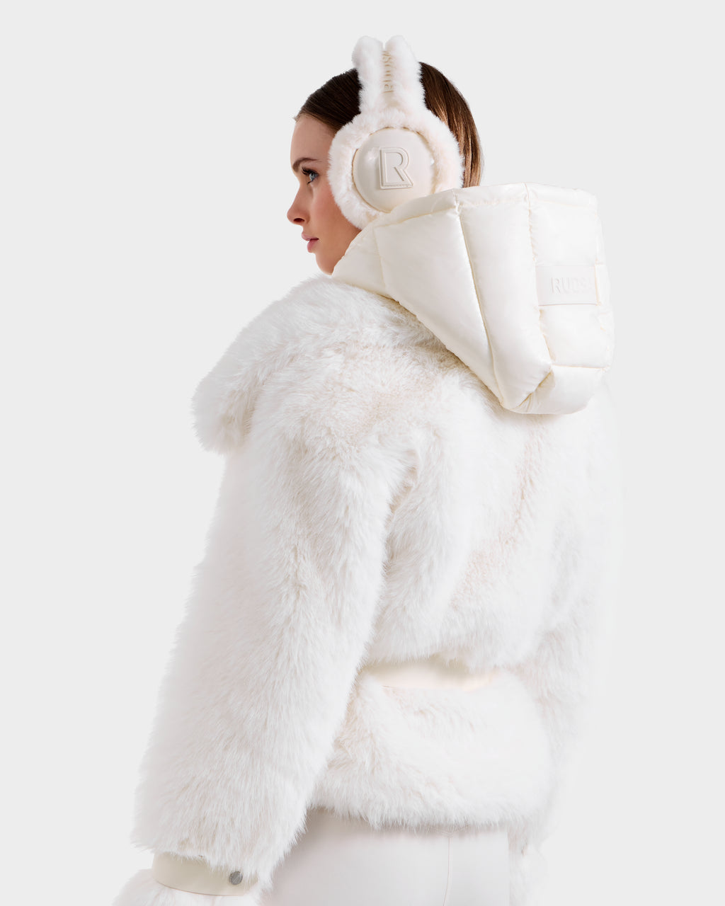 A woman models the CHLOE - 8125619 Iceland white faux fur jacket by Rudsak, paired with matching "R" earmuffs, viewed from the back against a plain light backdrop—an elegant take on winter fashion.