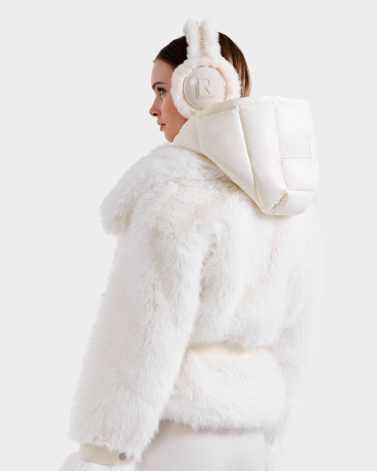 A woman models the CHLOE - 8125619 Iceland white faux fur jacket by Rudsak, paired with matching "R" earmuffs, viewed from the back against a plain light backdrop—an elegant take on winter fashion.