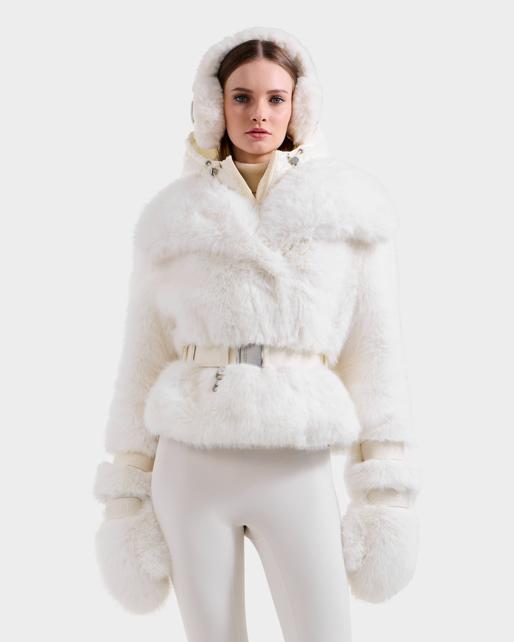 A woman models the Rudsak CHLOE - 8125619 Iceland women’s faux fur jacket with hood, matching belt, and large mittens, styled with white fitted pants—a must-have piece for your winter wardrobe.