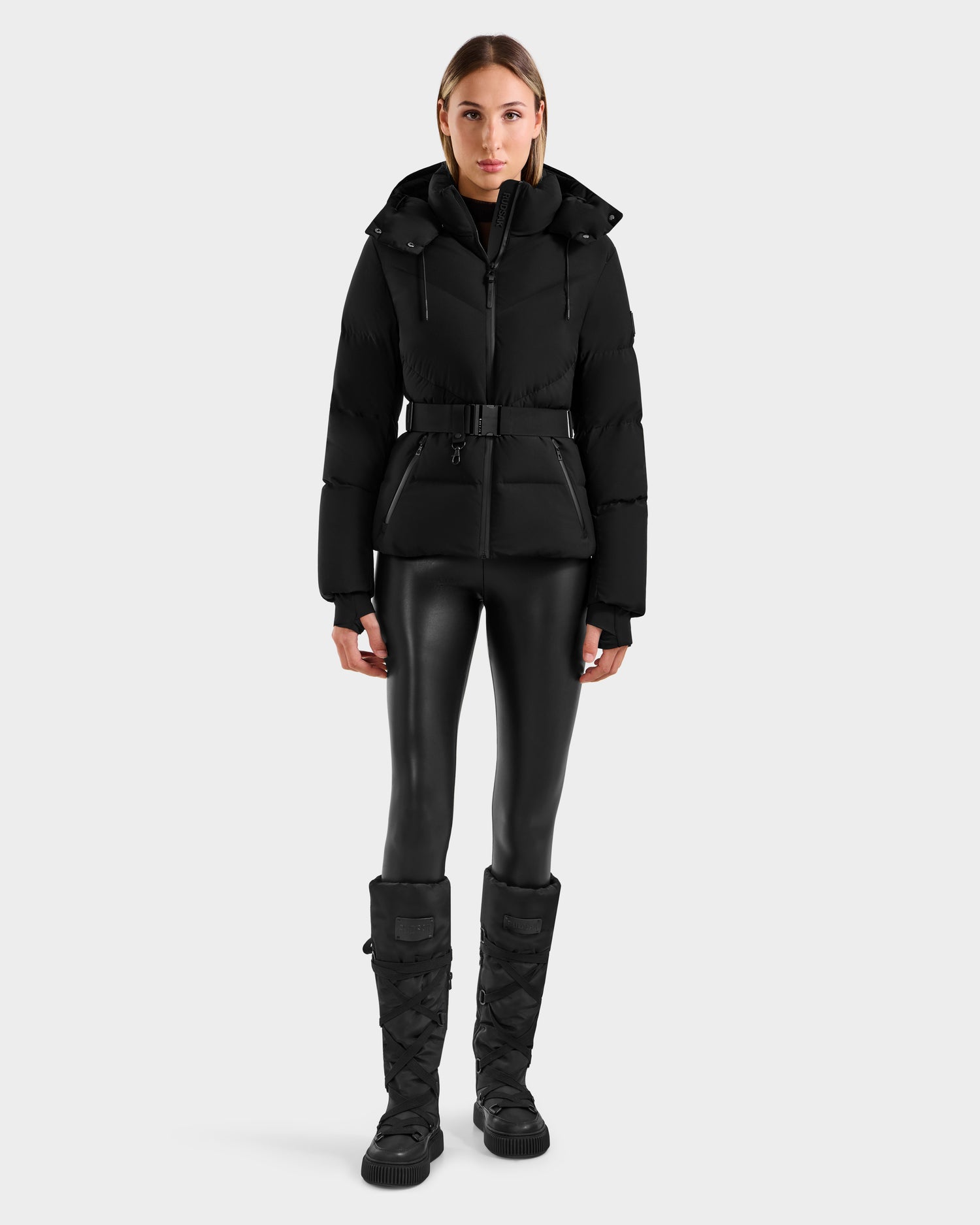A woman faces forward wearing the Rudsak CHRISTELLE M - L125542 Black quilted down puffer jacket with a belt, paired with black shiny leggings and knee-high boots against a plain light gray background.