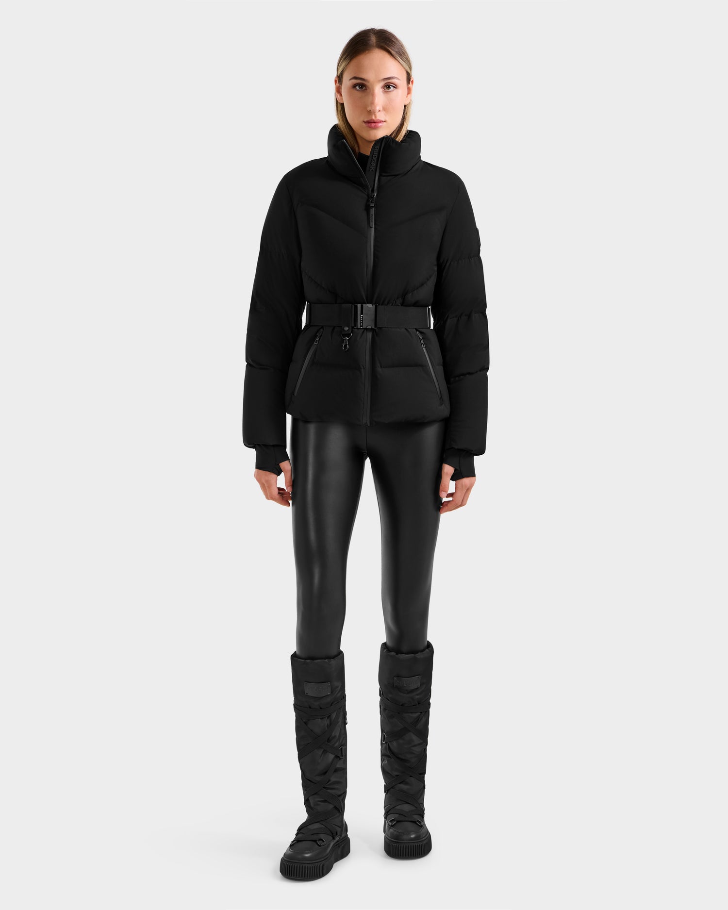 A woman stands facing forward in the Rudsak CHRISTELLE M - L125542 Black belted quilted down puffer jacket against a plain white background.