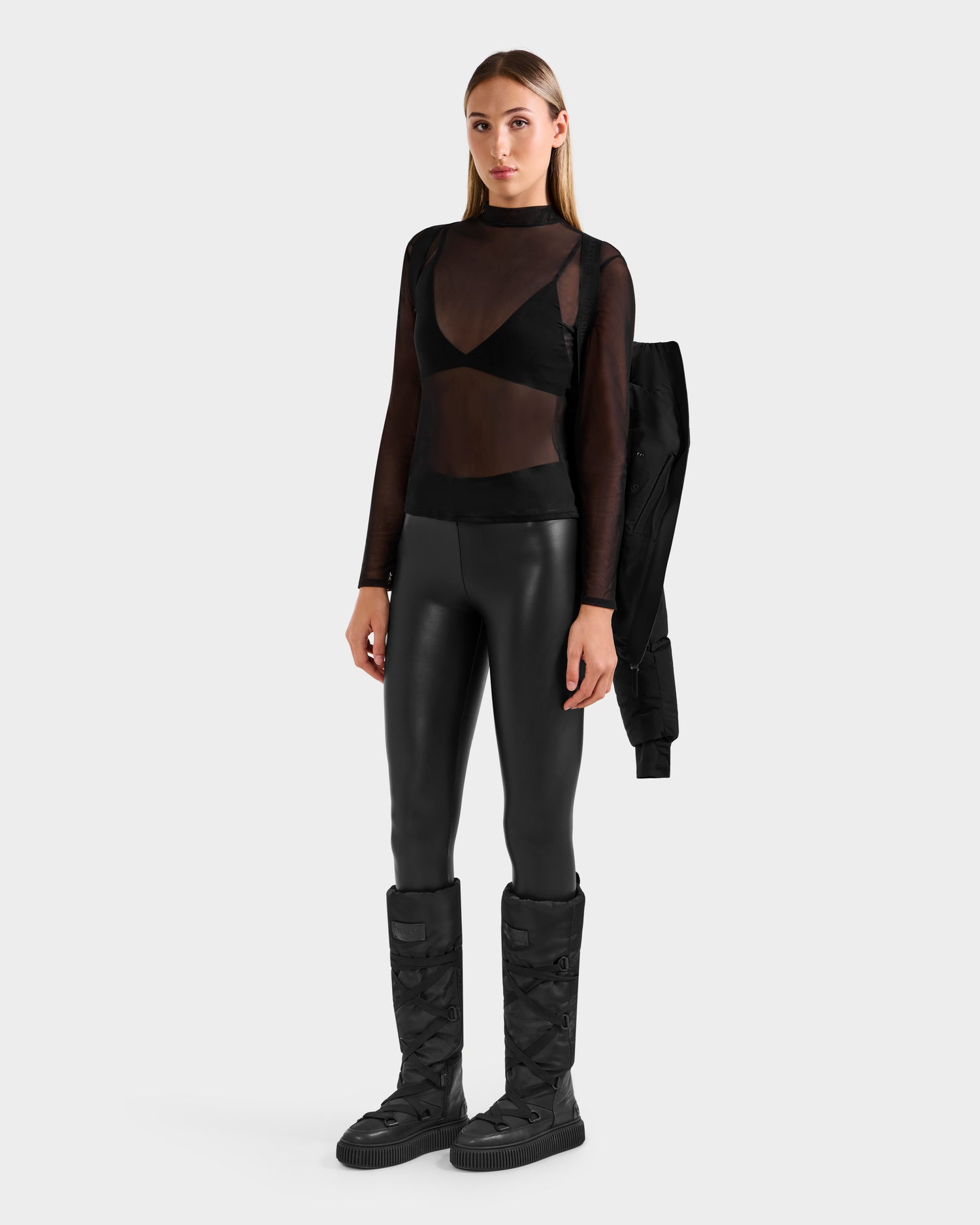 A woman with straight light brown hair and a neutral expression wears the Rudsak CHRISTELLE M - L125542 Black weather-resistant recycled shell jacket draped over one shoulder, styled with a sheer black top, black bra, faux leather pants, and large boots.