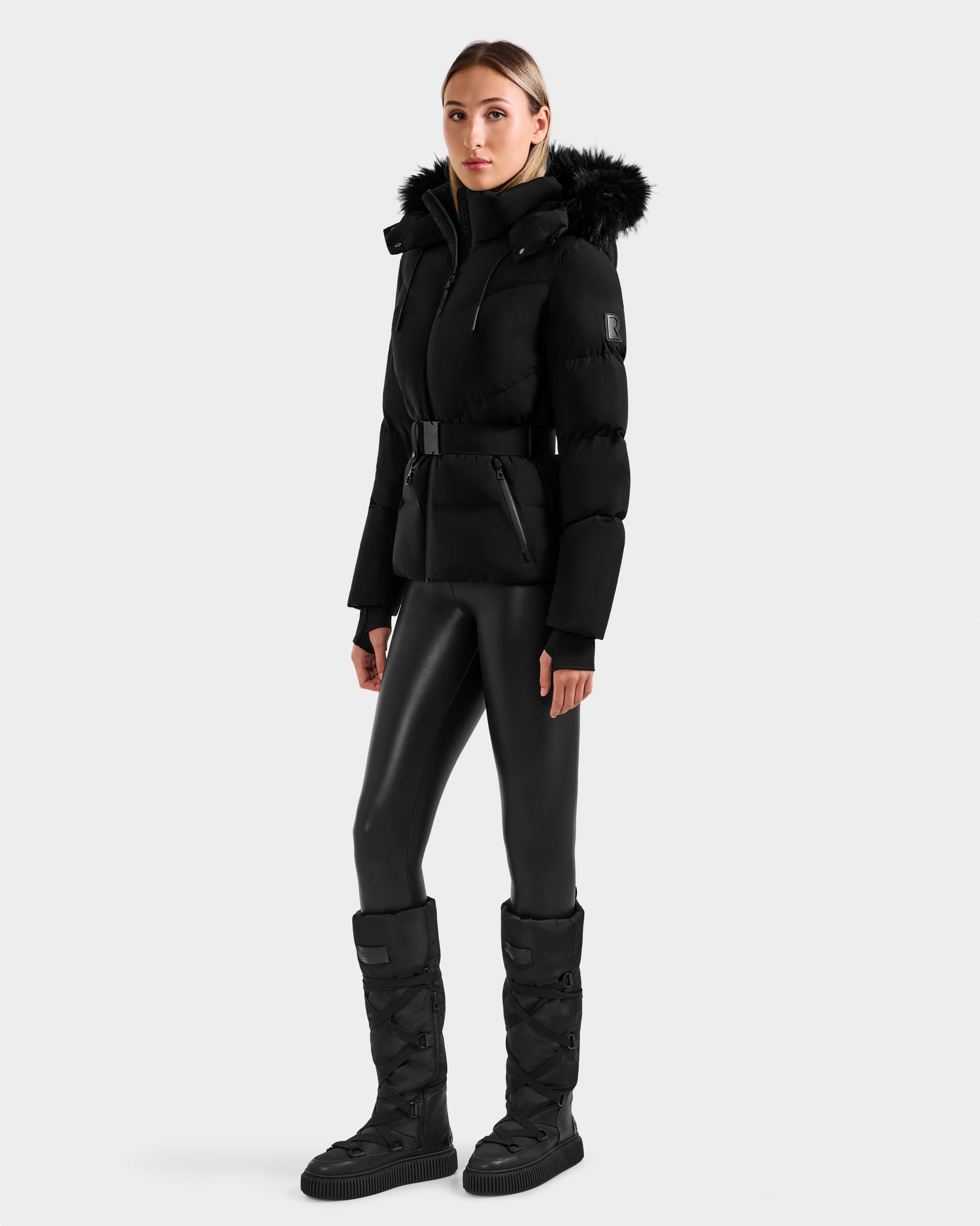 A woman stands against a plain background, wearing Rudsak's CHRISTELLE M - L125542 Black quilted down puffer jacket with a fur-lined hood and belt, paired with black leggings and tall weather-resistant recycled shell puffer boots.