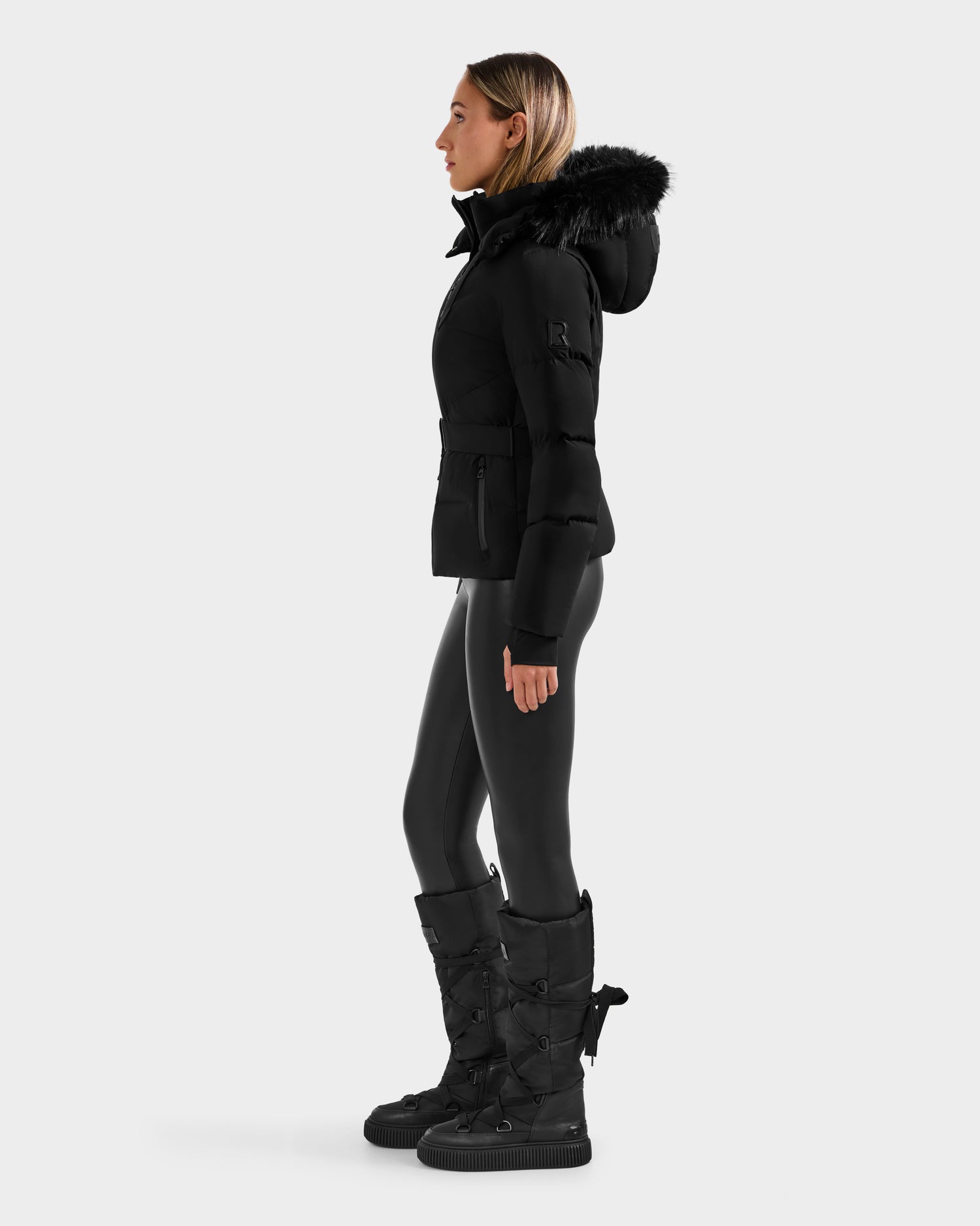 A woman stands in profile against a plain light background, wearing the Rudsak CHRISTELLE M - L125542 Black belted winter jacket with a fur-trimmed hood and RDS-certified down, black leggings, and tall black boots.