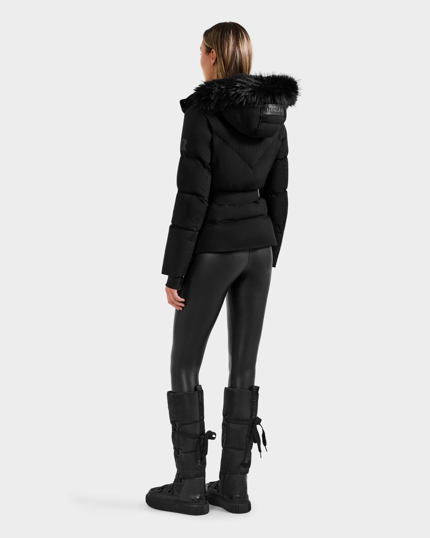 A woman, facing away, wears the Rudsak CHRISTELLE M - L125542 Black quilted down puffer jacket with a fur-lined hood, paired with black shiny leggings and knee-high lace-up boots. The background is plain light gray.