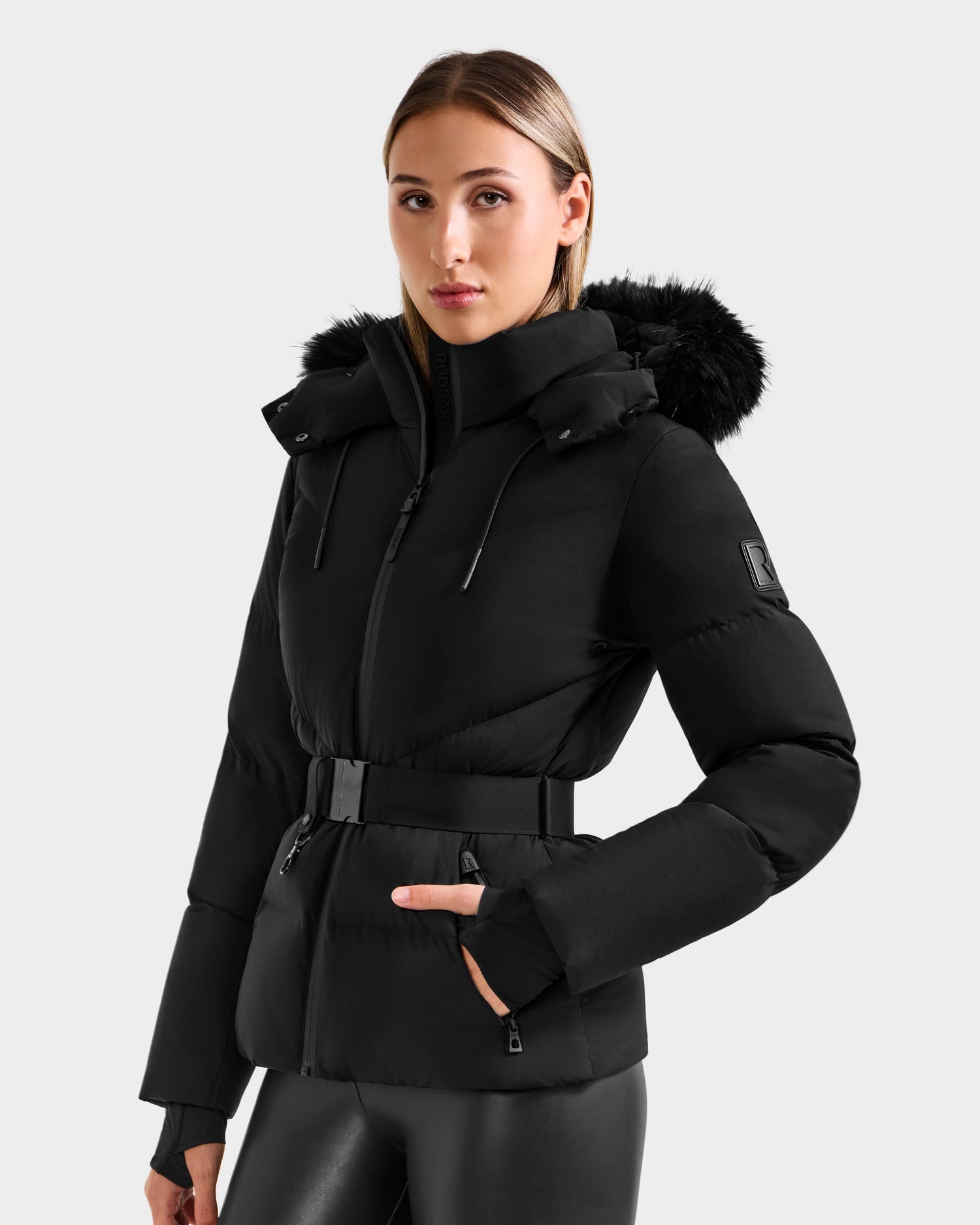 A woman wears the Rudsak CHRISTELLE M - L125542 Black quilted down puffer jacket with a fur-trimmed hood and black fitted pants, standing against a plain light gray background.