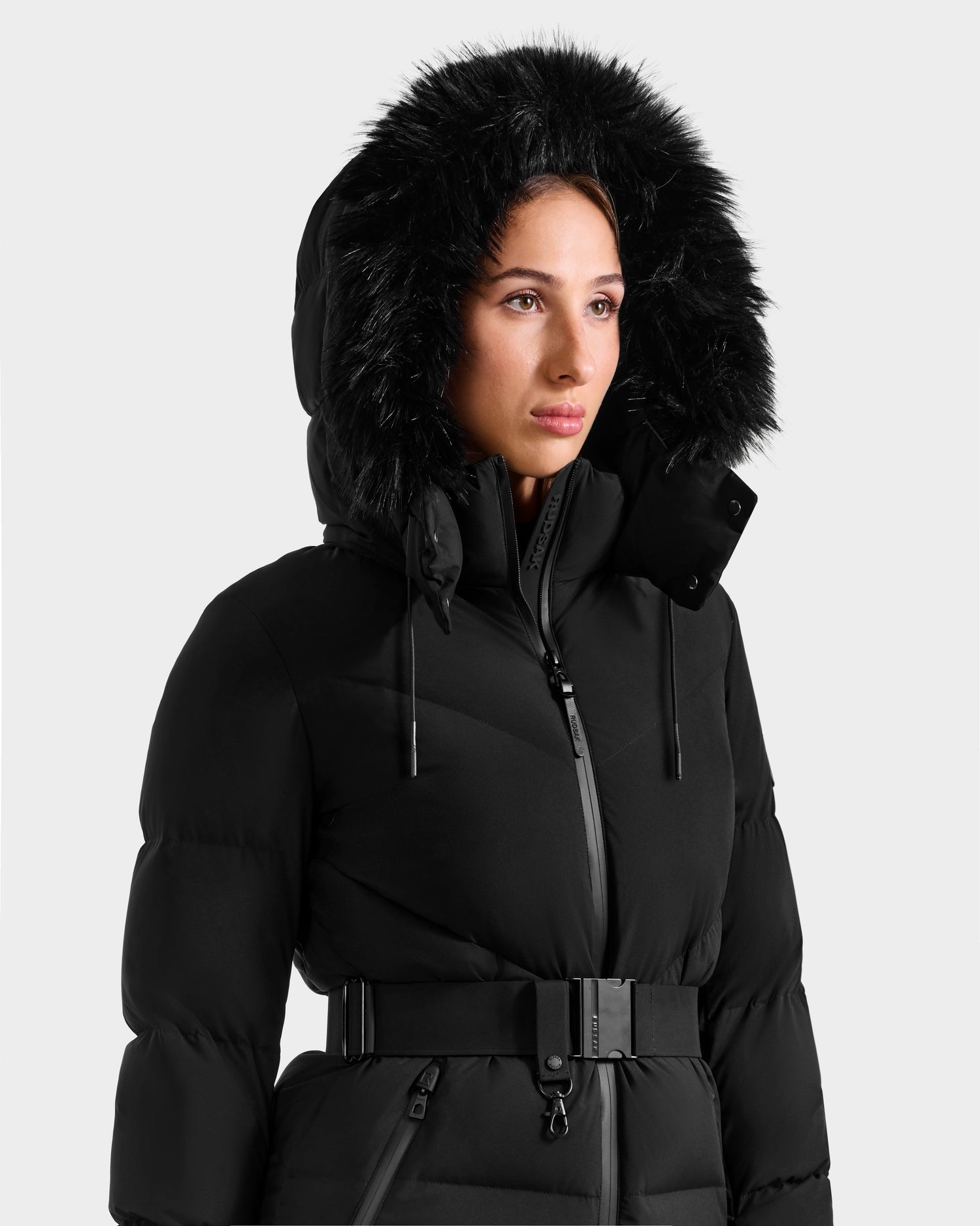 A person wears the Rudsak CHRISTELLE M - L125542 Black belted quilted down puffer jacket with a large black faux fur-trimmed hood, posing against a plain light background.