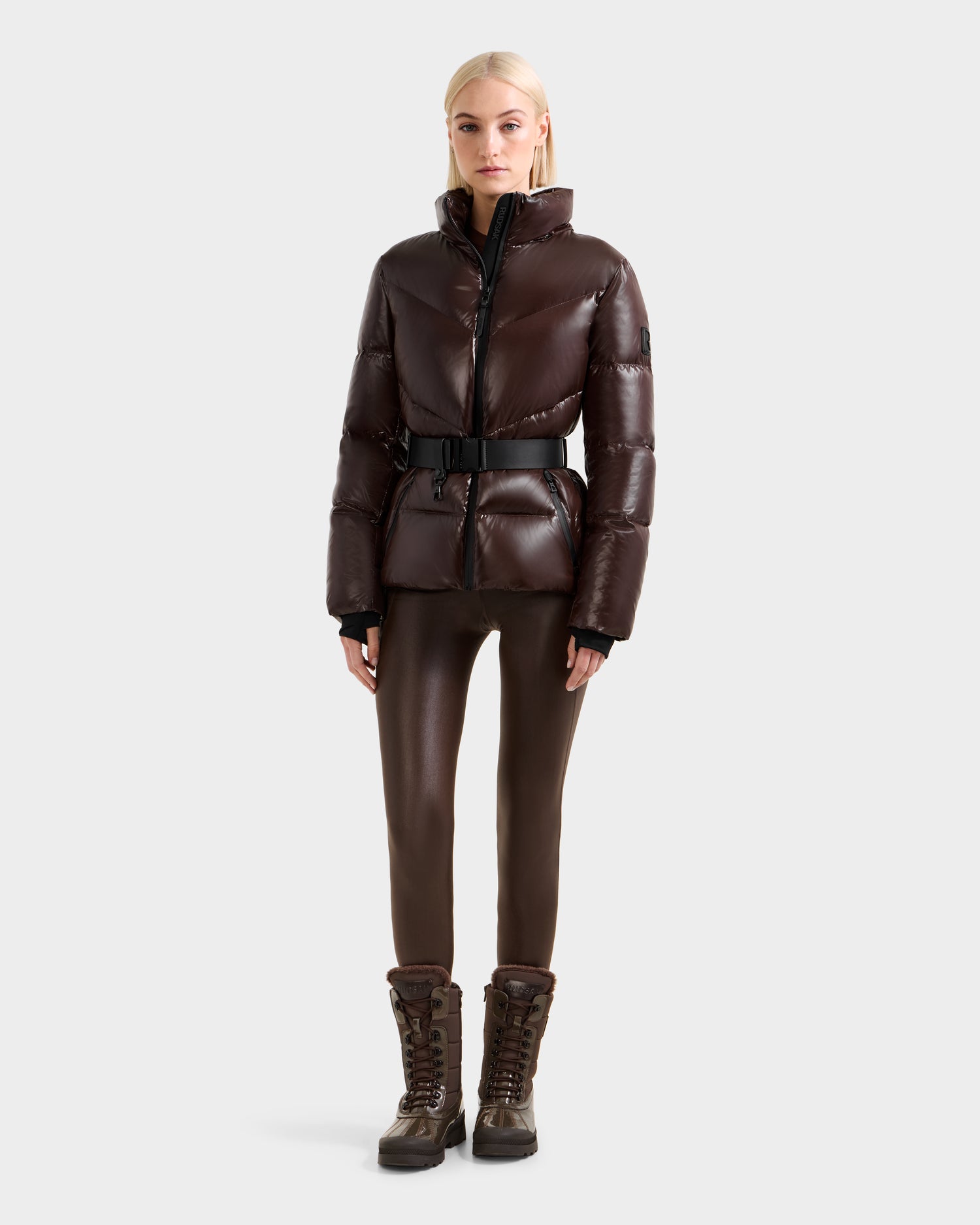 A woman wearing the Rudsak CHRISTELLE S - L125544 Espresso shiny dark brown down puffer jacket with a black belt, matching brown fitted pants, and lace-up winter boots stands facing forward against a light gray background.