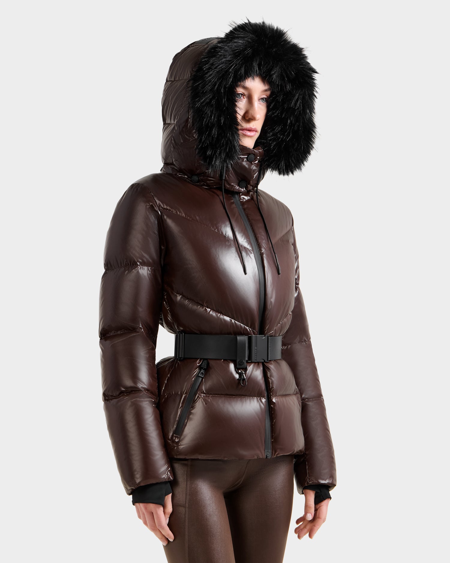 A woman wears the Rudsak CHRISTELLE S - L125544 Espresso down puffer jacket with a belted waist, front zipper, and hood with black faux fur trim. She stands against a plain light background, looking slightly to the side.