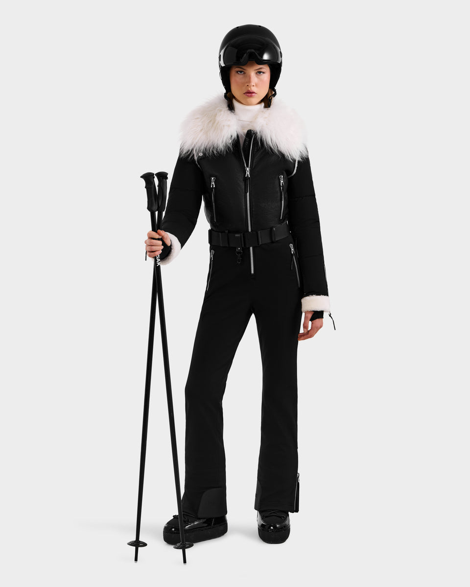 A woman in the Rudsak CHYNTIA - 8125611 Black ski outfit with a white fur collar, black helmet, and goggles holds black ski poles against a plain background. Pre-order now for shipping by November 30th.