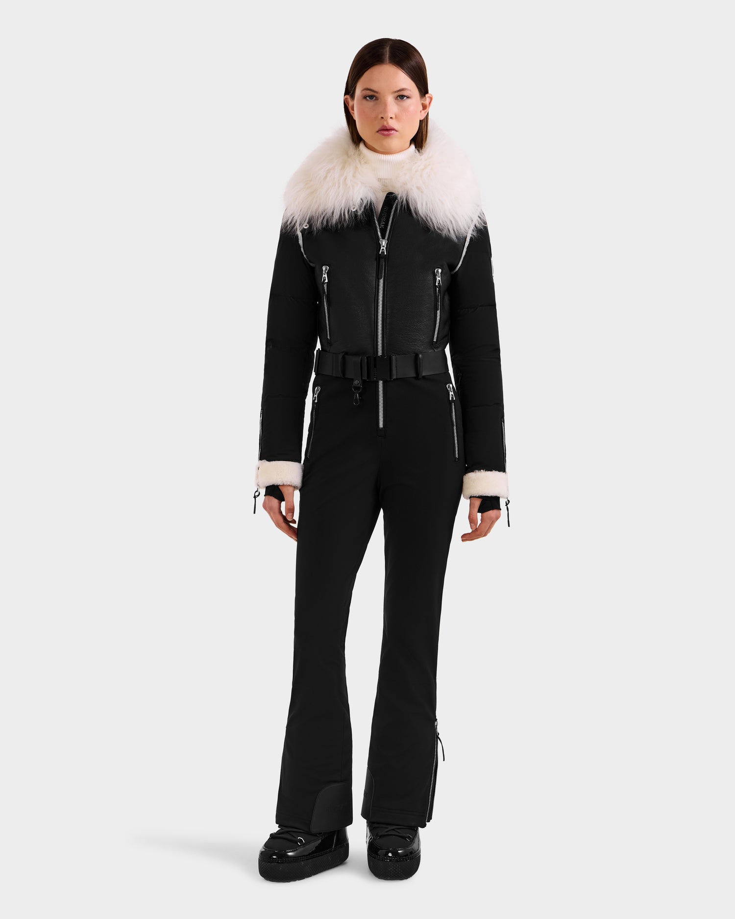 A woman faces forward in the Rudsak CHYNTIA - 8125611 Black ski suit, featuring a fitted silhouette, wide white faux fur collar and cuffs, black belt, multiple zippers, and black boots. Available for pre-order; ships by November 30th.