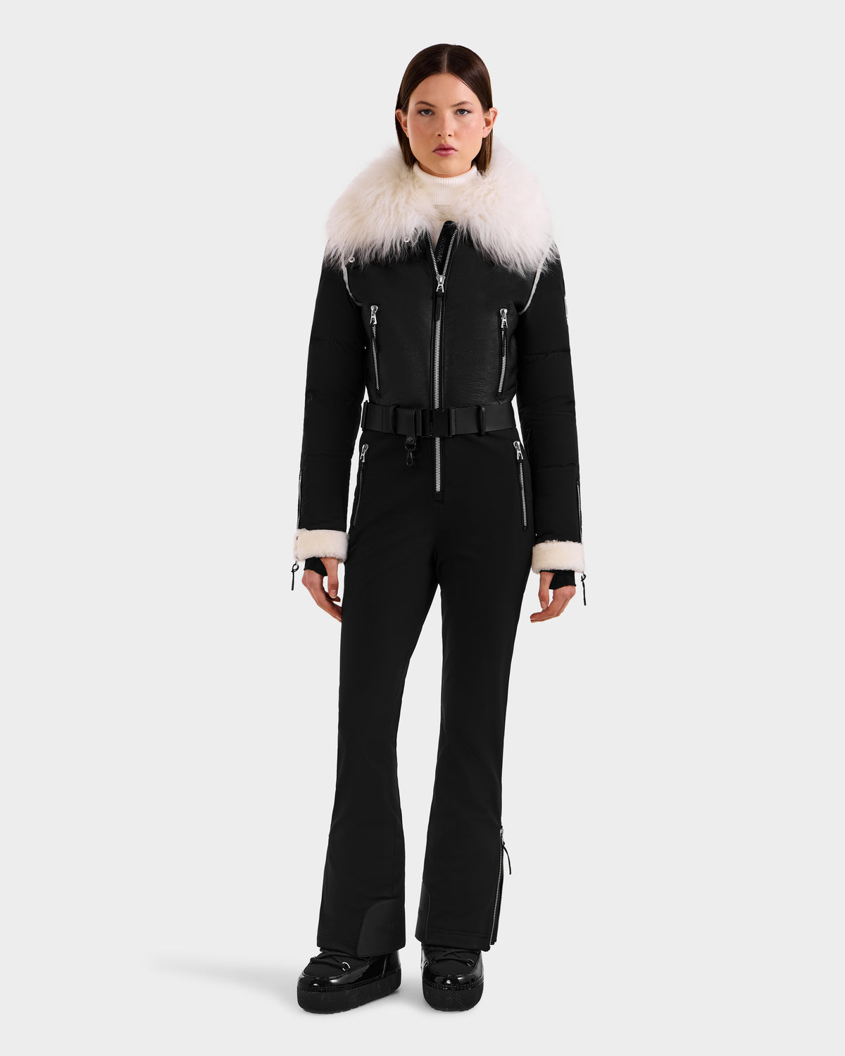 A woman faces forward in the Rudsak CHYNTIA - 8125611 Black ski suit, featuring a fitted silhouette, wide white faux fur collar and cuffs, black belt, multiple zippers, and black boots. Available for pre-order; ships by November 30th.