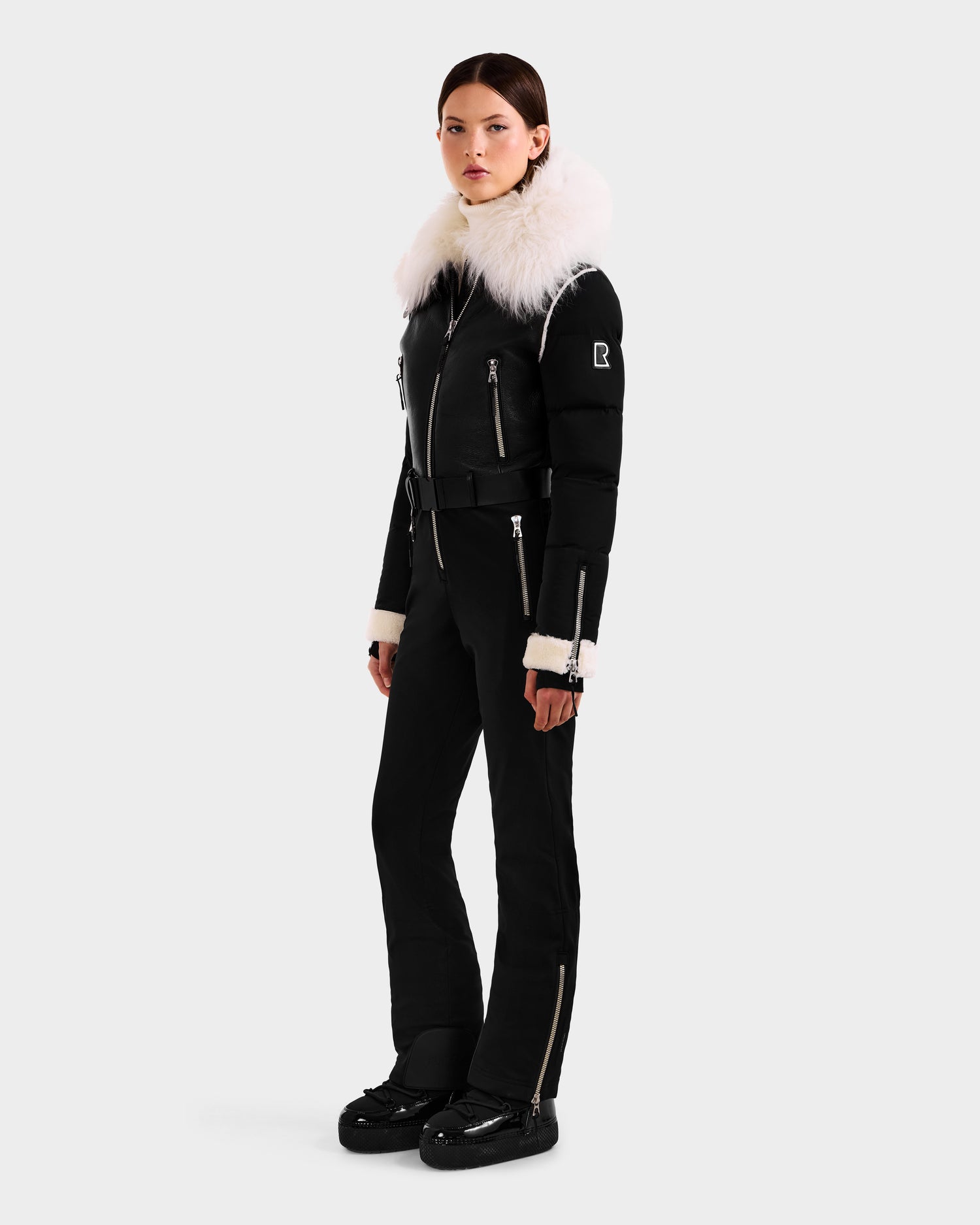 A woman wears the Rudsak CHYNTIA - 8125611 Black ski suit featuring a luxurious Mongolian-fur collar and cuffs, silver zippers, and black boots, set against a plain light gray background.