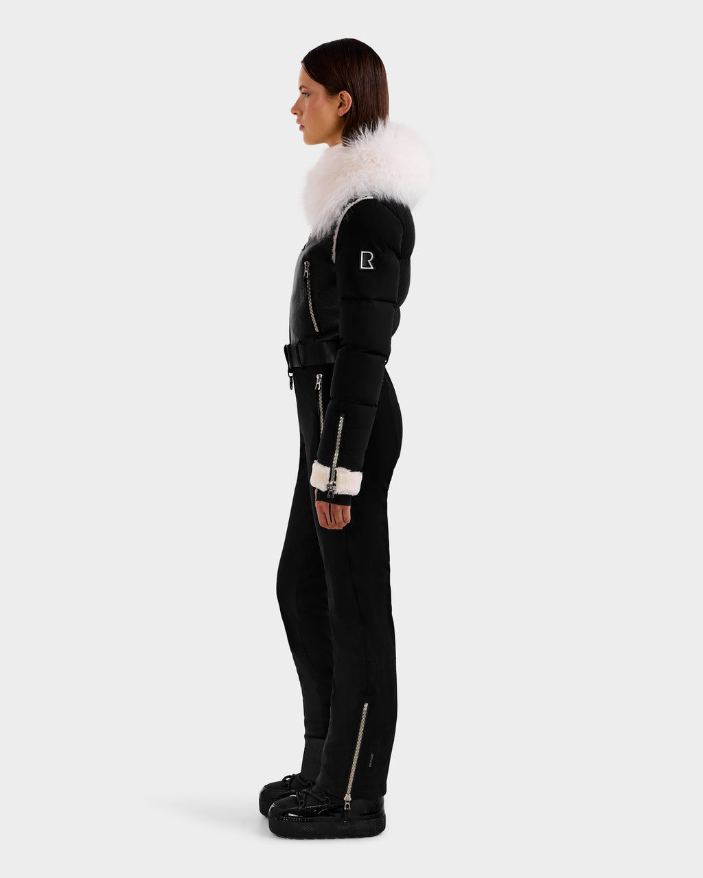 A woman in profile wears the Rudsak CHYNTIA - 8125611 Black ski suit, featuring a fitted silhouette, large white Mongolian-fur collar, black boots, and white cuffs against a plain light gray background.