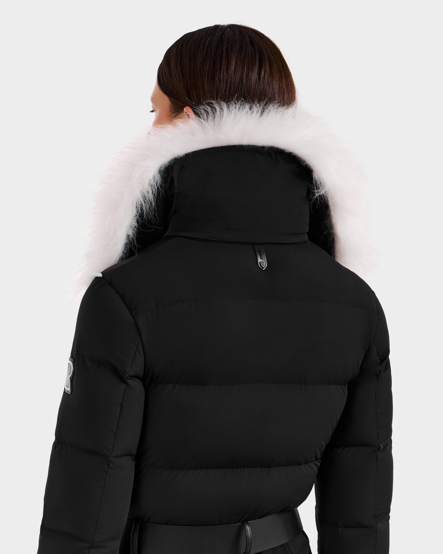 A person with straight brown hair is seen from behind, wearing the Rudsak CHYNTIA - 8125611 Black puffer jacket with a Mongolian-fur collar, sleeve patch, black waist belt, and RECCO rescue technology.