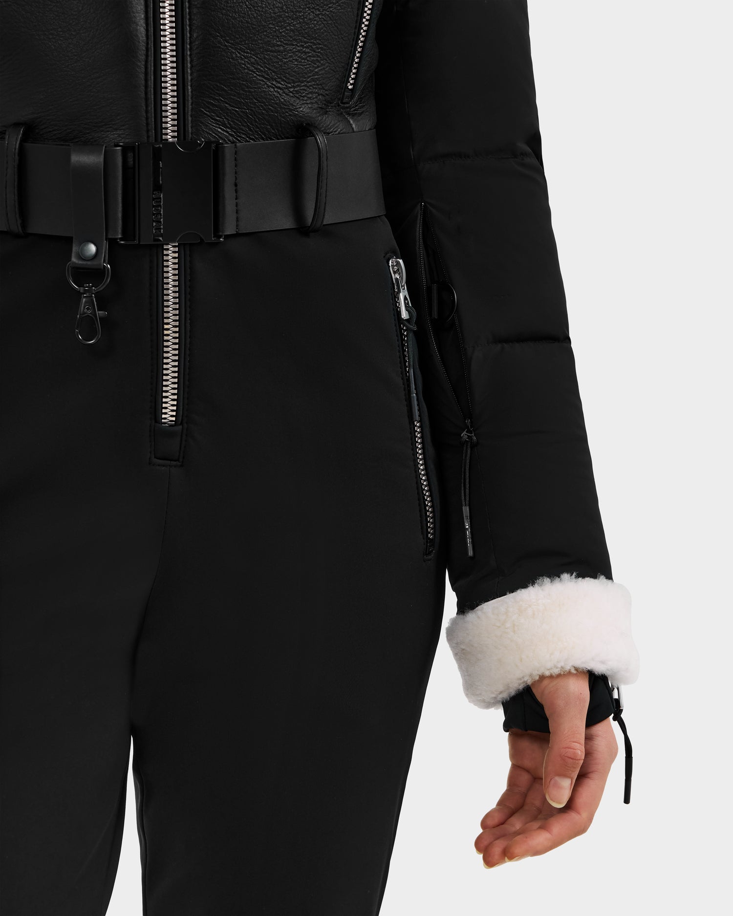 A person wears the Rudsak CHYNTIA - 8125611 Black jacket, featuring a zip pocket, belted waist, Mongolian-fur collar, and cream shearling cuffs with black pants. Only the lower torso, right arm, and hand are visible against a plain background.