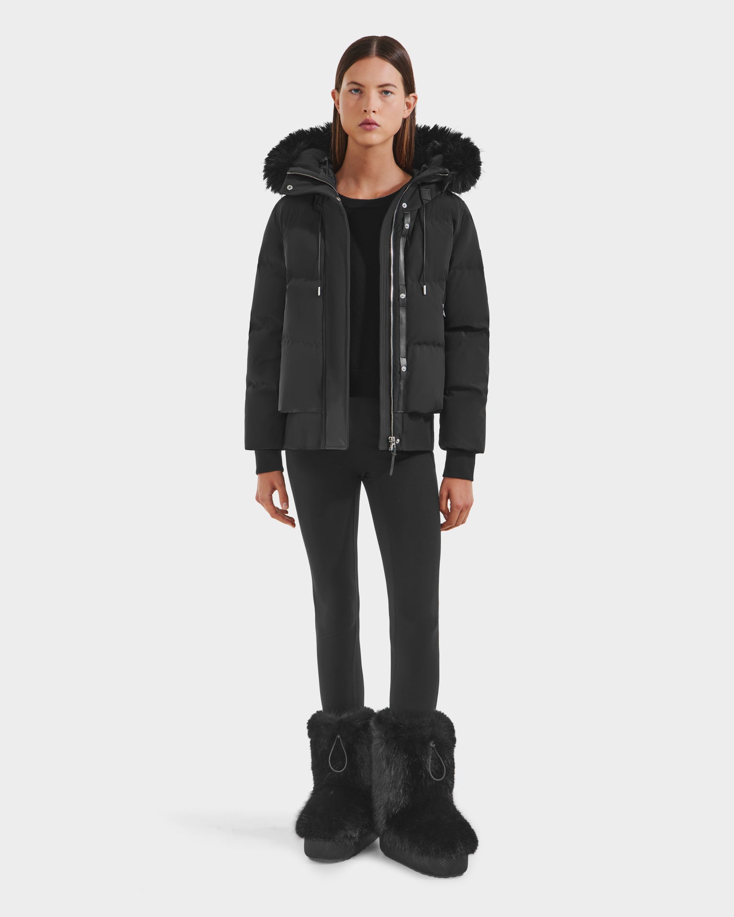 A woman faces forward in the Rudsak CIEYNA - 8125579 Black down-filled bomber with a fur-trimmed hood, paired with black leggings and fur boots—highlighting this winter essential against a plain white background.