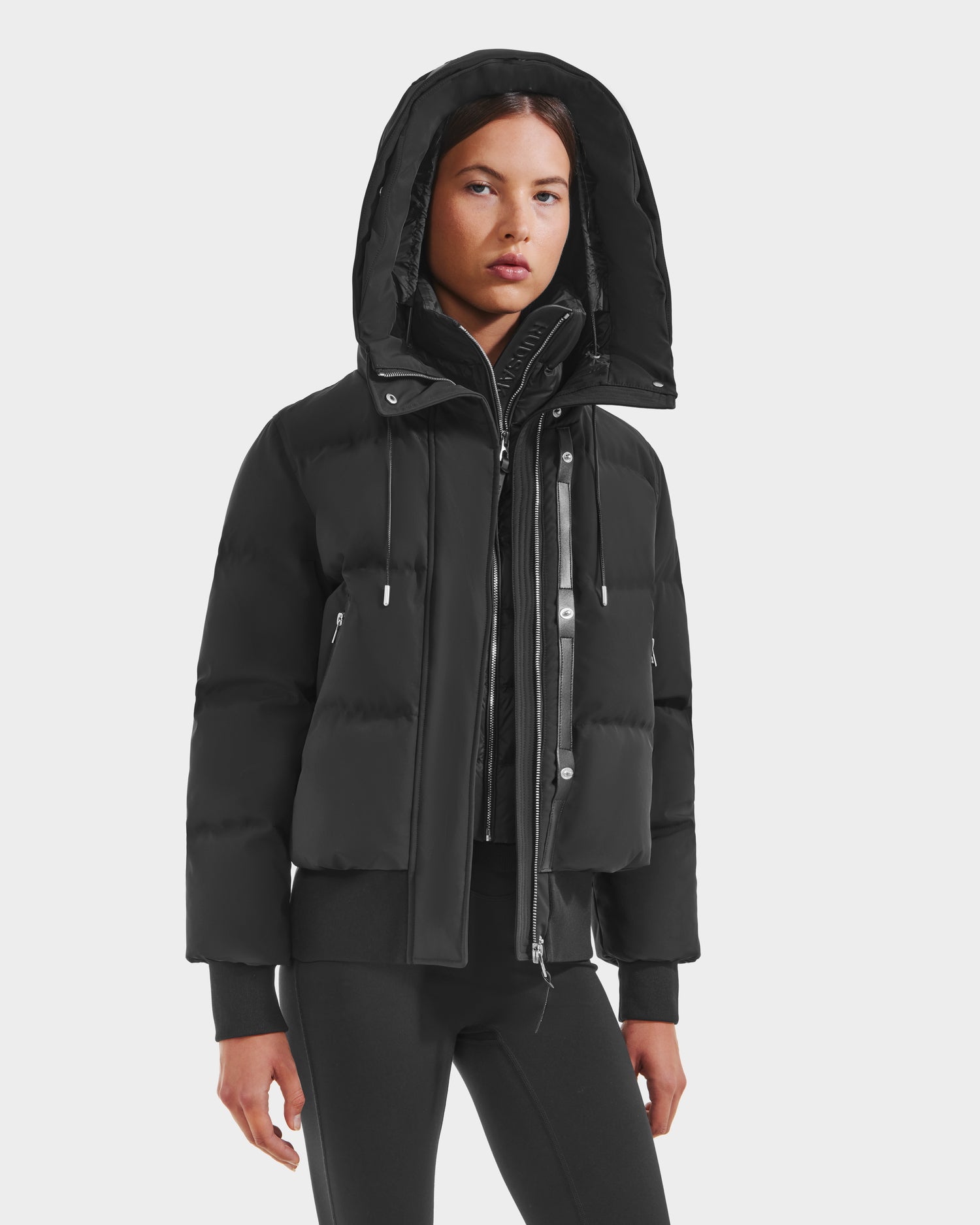 A woman models the Rudsak CIEYNA - 8125579 Black puffer jacket over a zipped top and fitted black pants, standing against a plain white background with a neutral expression.