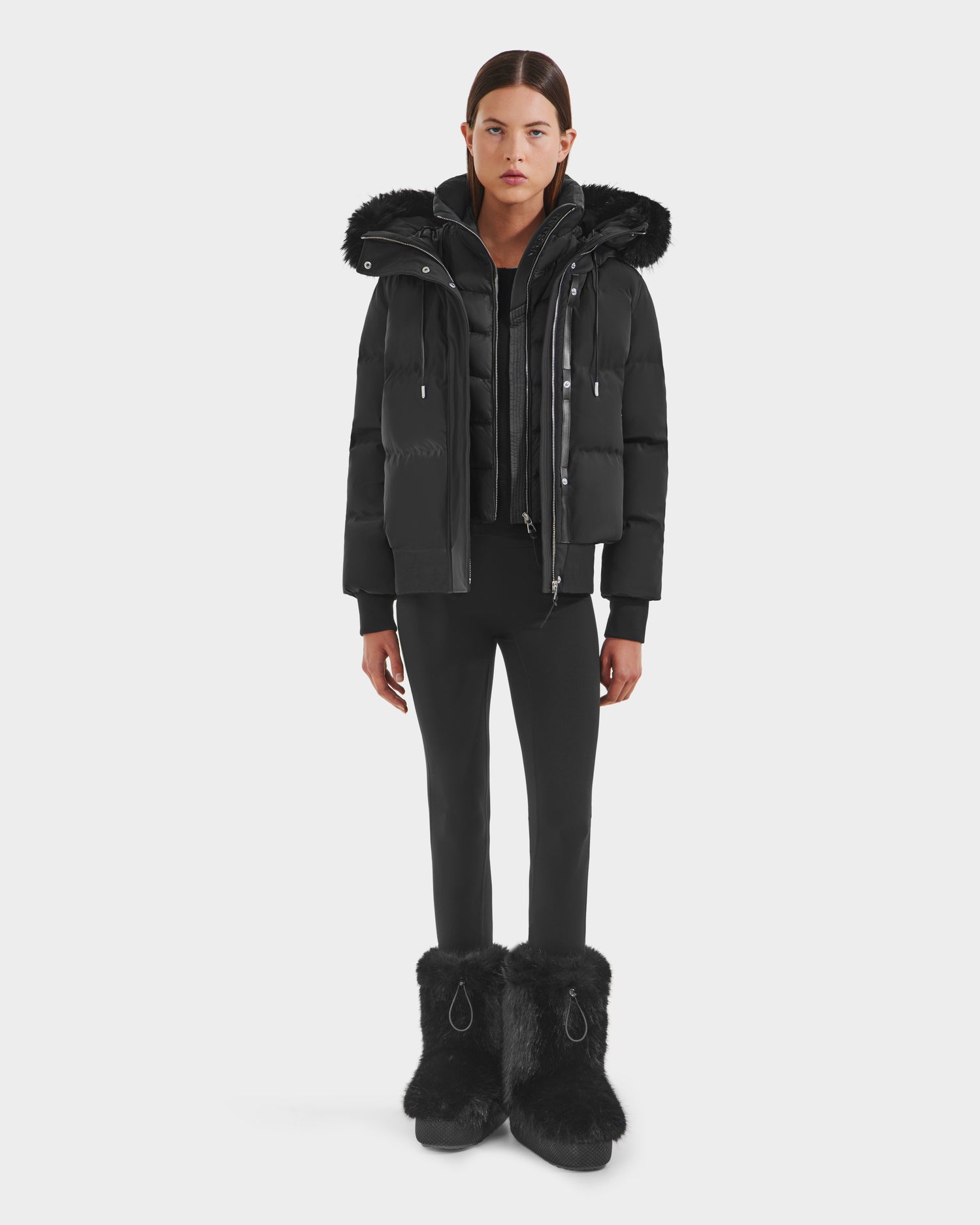 A person stands facing forward in the Rudsak CIEYNA - 8125579 Black weather-resistant puffer jacket with a hood, paired with black pants and furry boots, set against a plain white background—a winter essential.