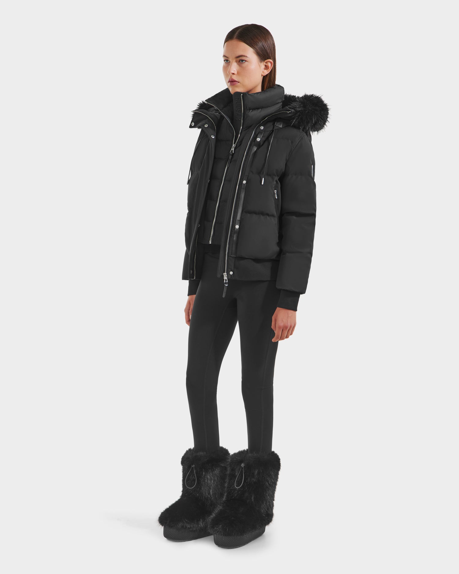 A woman models the Rudsak CIEYNA - 8125579 Black jacket with a fur-trimmed hood, styled with a black zip-up top, fitted black pants, and large furry boots against a plain white background.