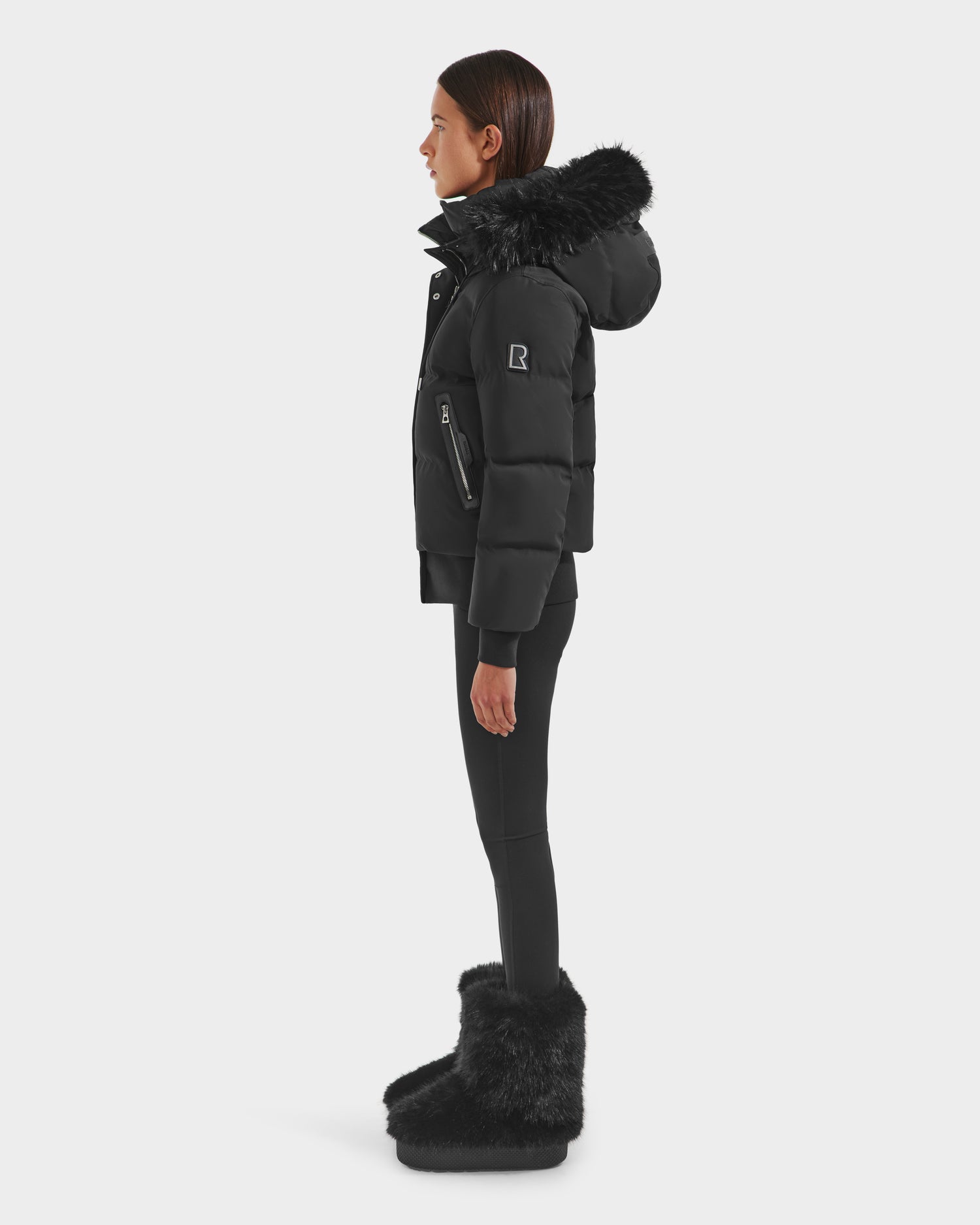 A woman stands in profile against a plain white background wearing the Rudsak CIEYNA - 8125579 Black weather-resistant jacket with a fur-lined hood, paired with black leggings and furry black boots.