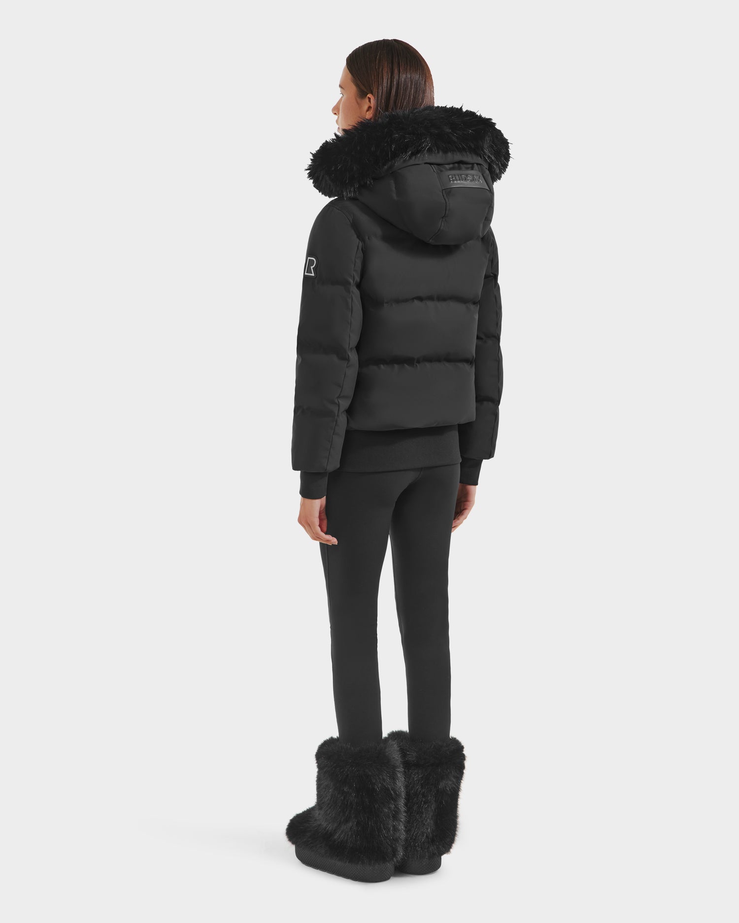 A woman faces away, wearing the Rudsak CIEYNA - 8125579 Black jacket with a fur-lined hood, paired with black leggings and fur boots—modeling this winter essential against a plain white background.
