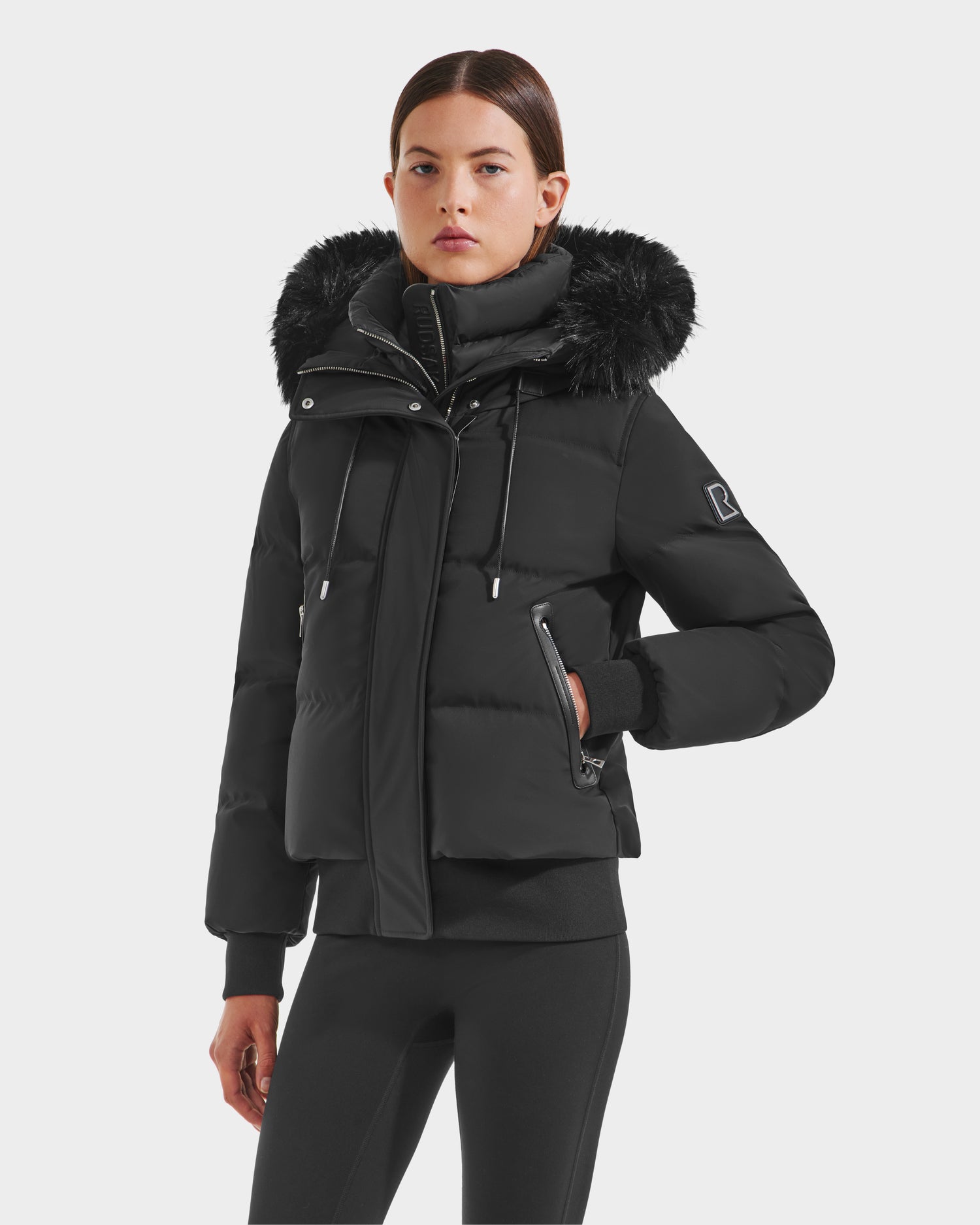 A woman wears the Rudsak CIEYNA - 8125579 Black puffer jacket with faux fur hood trim, zippered front and pockets, recycled weather-resistant shell, and black fitted pants, standing out against a plain light background.