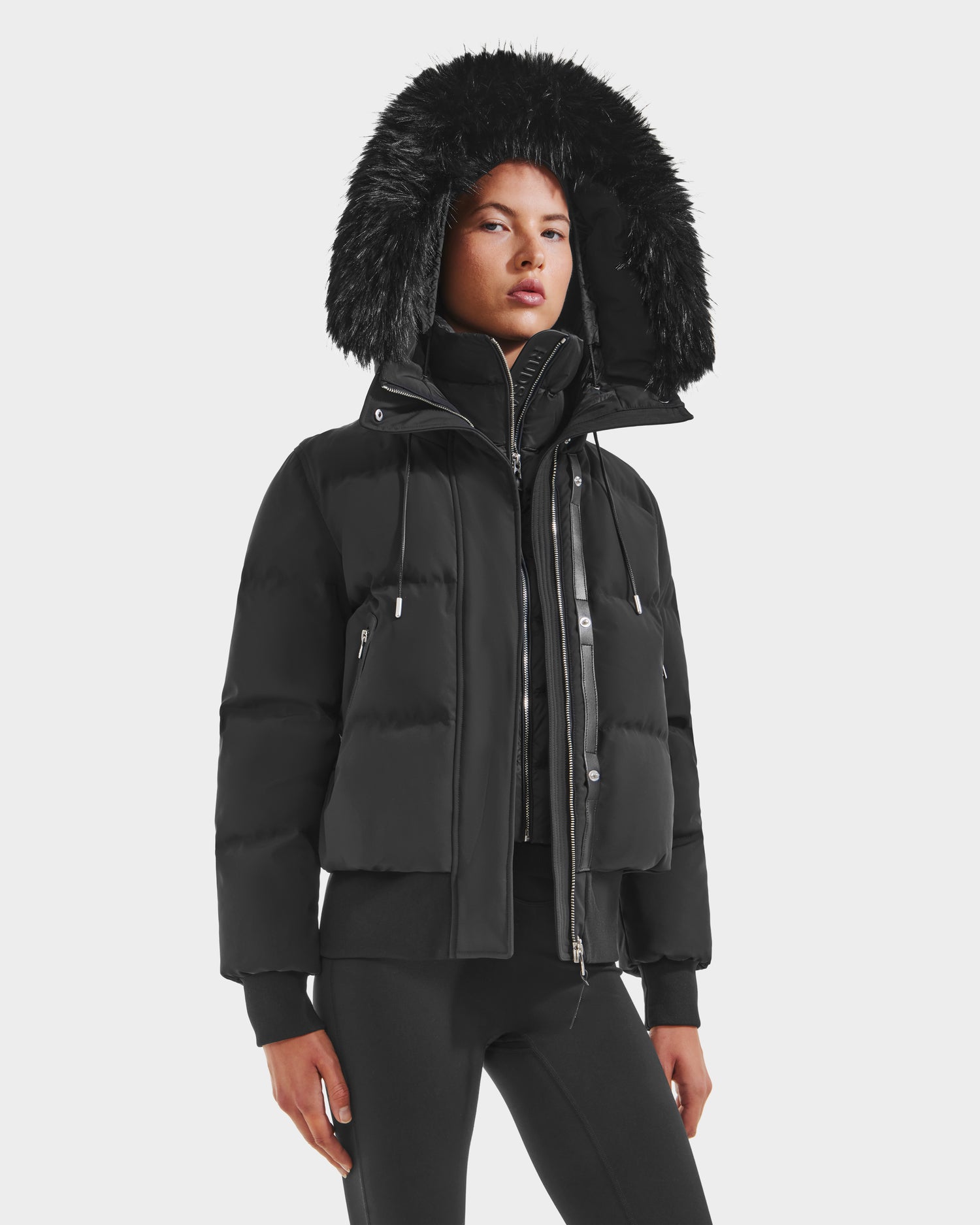 A woman stands against a white background in the Rudsak CIEYNA - 8125579 Black puffer jacket, featuring a weather-resistant zip-up front and large faux fur-trimmed hood, paired with black leggings for a chic winter look.