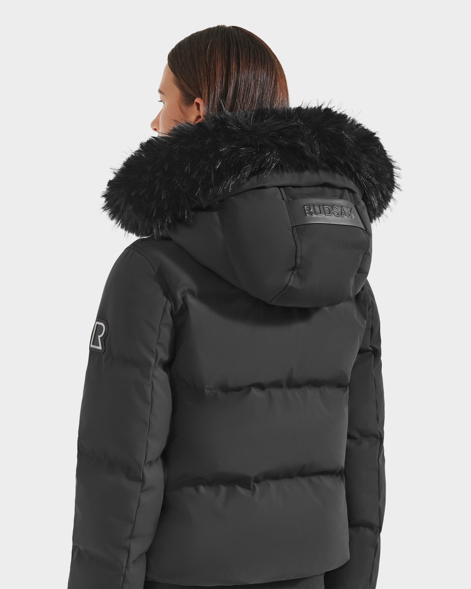 A person facing away wears the Rudsak CIEYNA - 8125579 Black down-filled bomber with a large furry hood and Rudsak logo on the back. Straight brown hair peeks above the collar—a true winter essential.