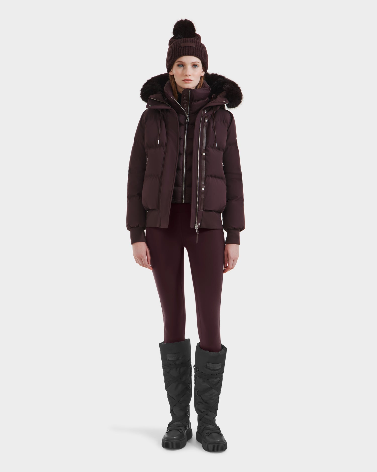 A woman stands facing forward against a white background, wearing the Rudsak CIEYNA - 8125579 Bordeaux down-filled bomber with faux fur hood.