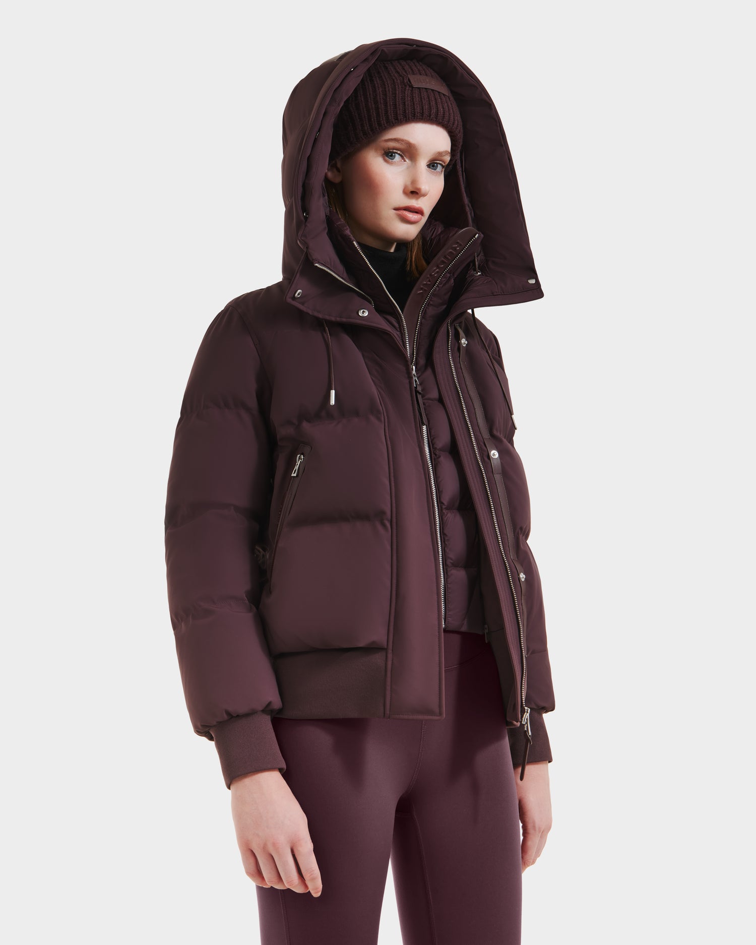 A woman stands against a plain white background wearing the Rudsak CIEYNA - 8125579 Bordeaux weather-resistant shell puffer jacket with hood, a matching beanie, and fitted leggings.