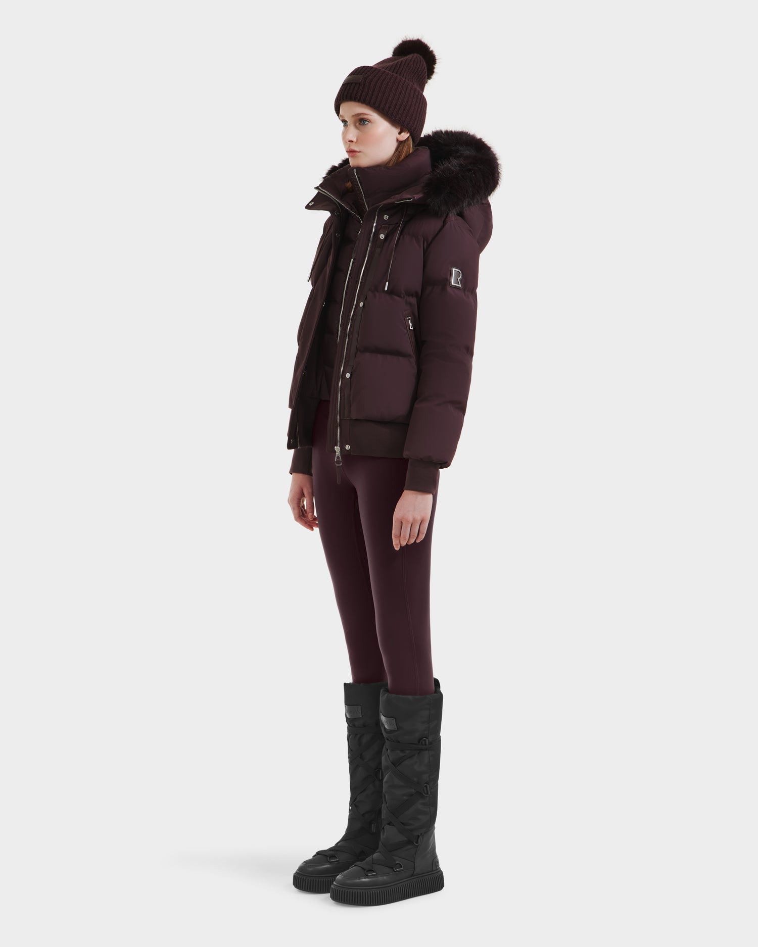 A woman in the Rudsak CIEYNA - 8125579 Bordeaux down-filled bomber with a faux fur hood, matching leggings, a knit beanie, and tall black winter boots stands facing left against a plain white background.