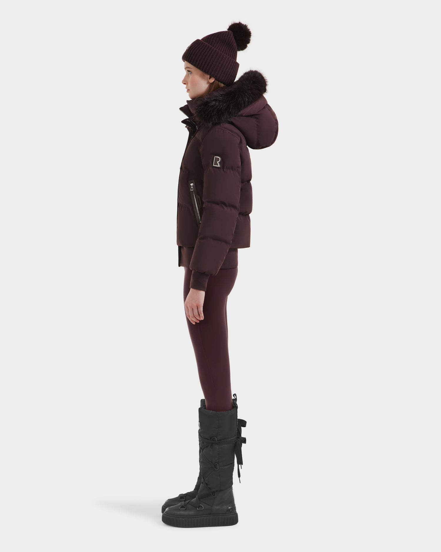 A person in profile wears the Rudsak CIEYNA - 8125579 Bordeaux weather-resistant puffer with faux fur hood, matching leggings, tall black boots, and a knit pom-pom beanie in deep burgundy and black.
