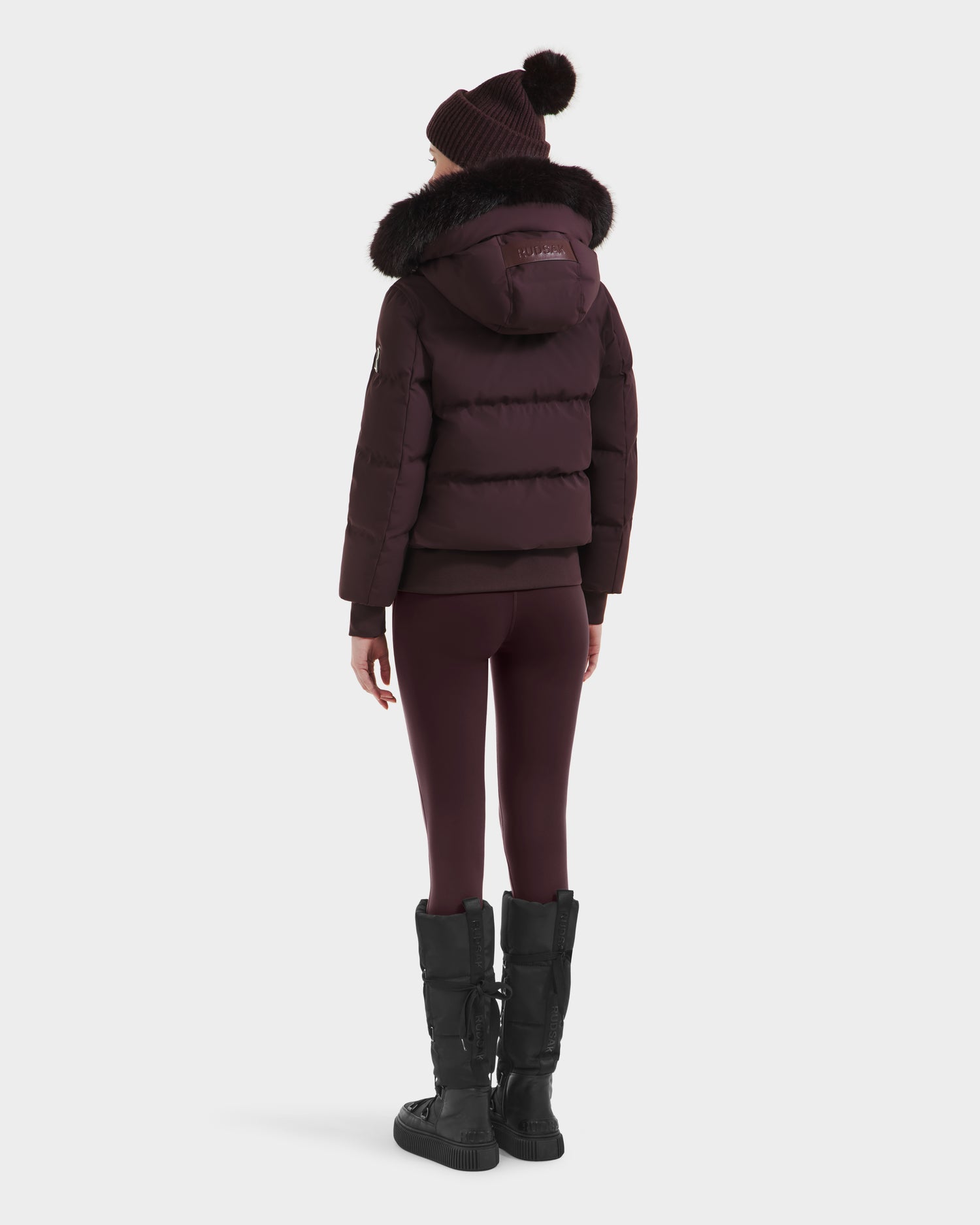 A person faces away from the camera, wearing Rudsaks CIEYNA - 8125579 Bordeaux down-filled bomber with faux fur hood, paired with matching leggings, a knit beanie, and black winter boots against a plain background.