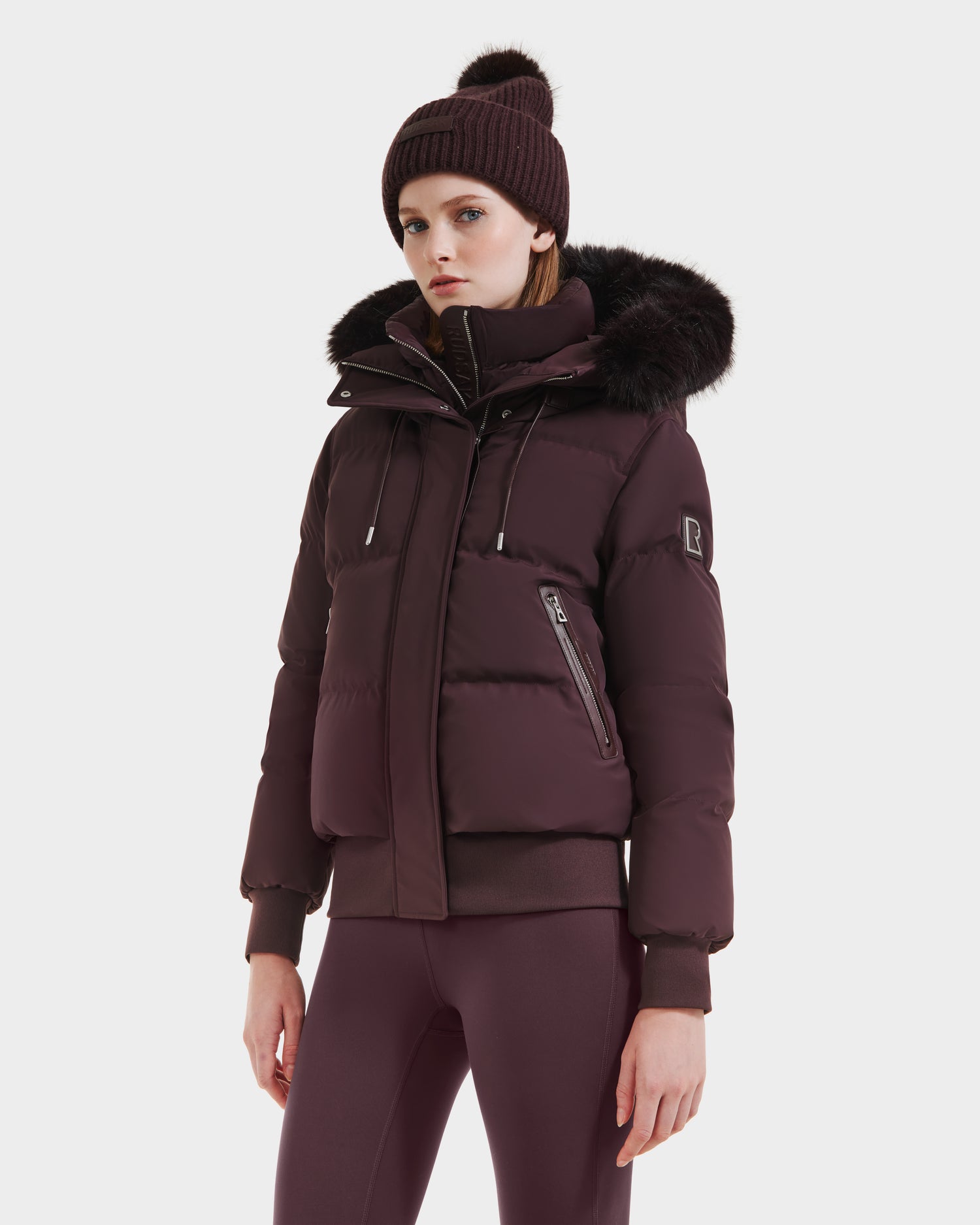 A woman stands against a white background wearing the Rudsak CIEYNA - 8125579 Bordeaux puffer jacket, made from recycled weather-resistant shell with a fur-trimmed hood, paired with maroon leggings and a knit beanie.