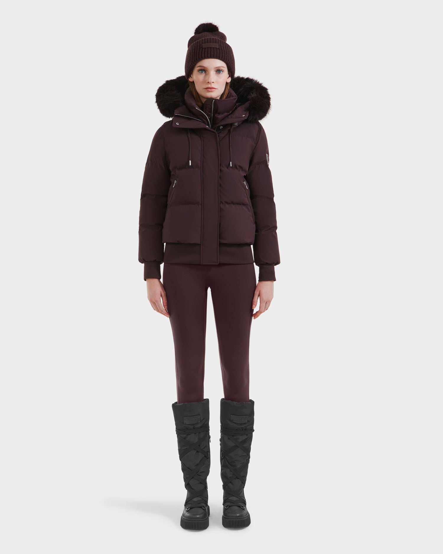 A person faces forward against a white background, wearing the Rudsak CIEYNA - 8125579 Bordeaux winter jacket with a faux fur hood, paired with matching leggings, a knit beanie, and tall black quilted boots.