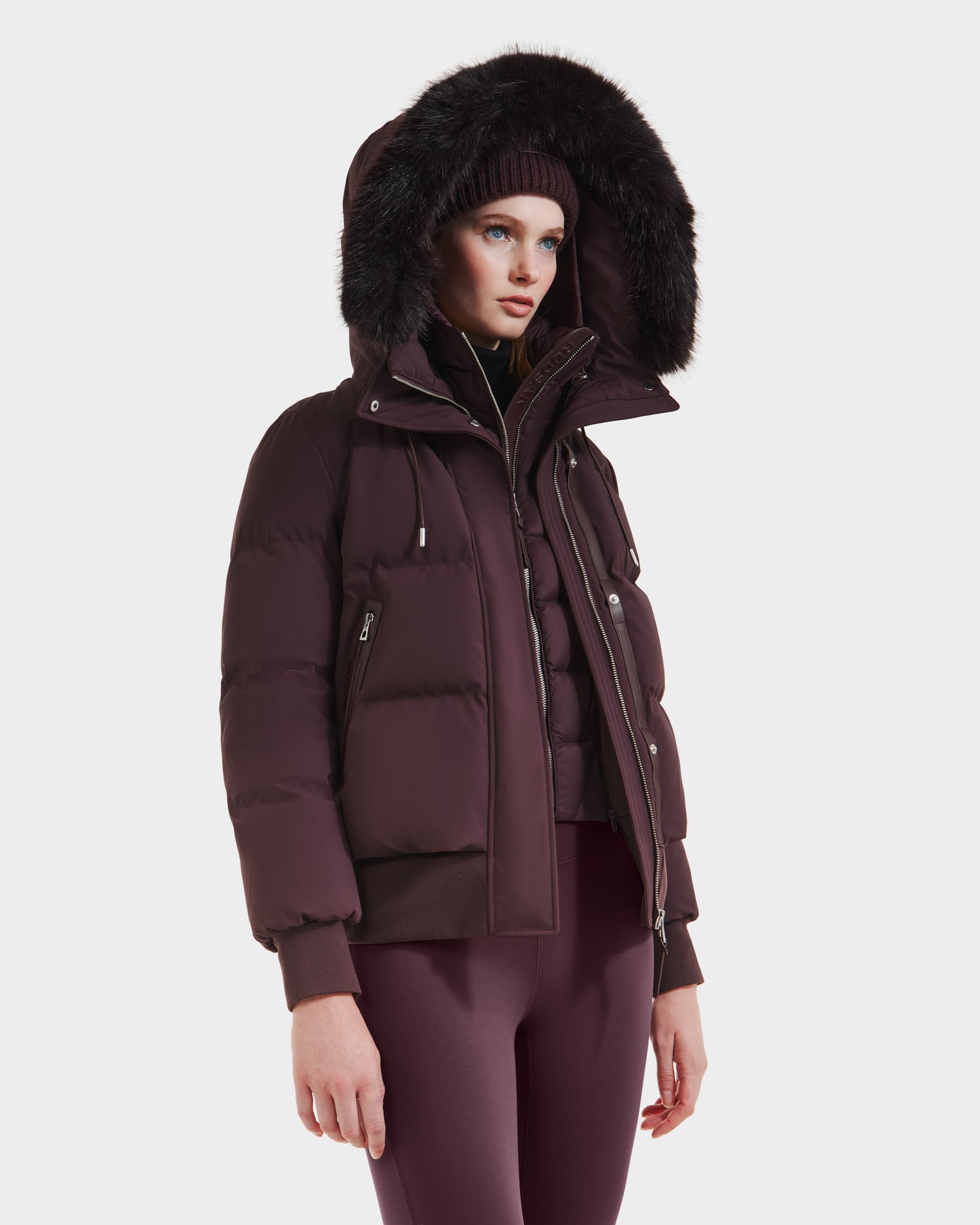A woman in the Rudsak CIEYNA - 8125579 Bordeaux weather-resistant puffer jacket with a fur-trimmed hood, maroon knit hat, and matching leggings stands against a plain white background.