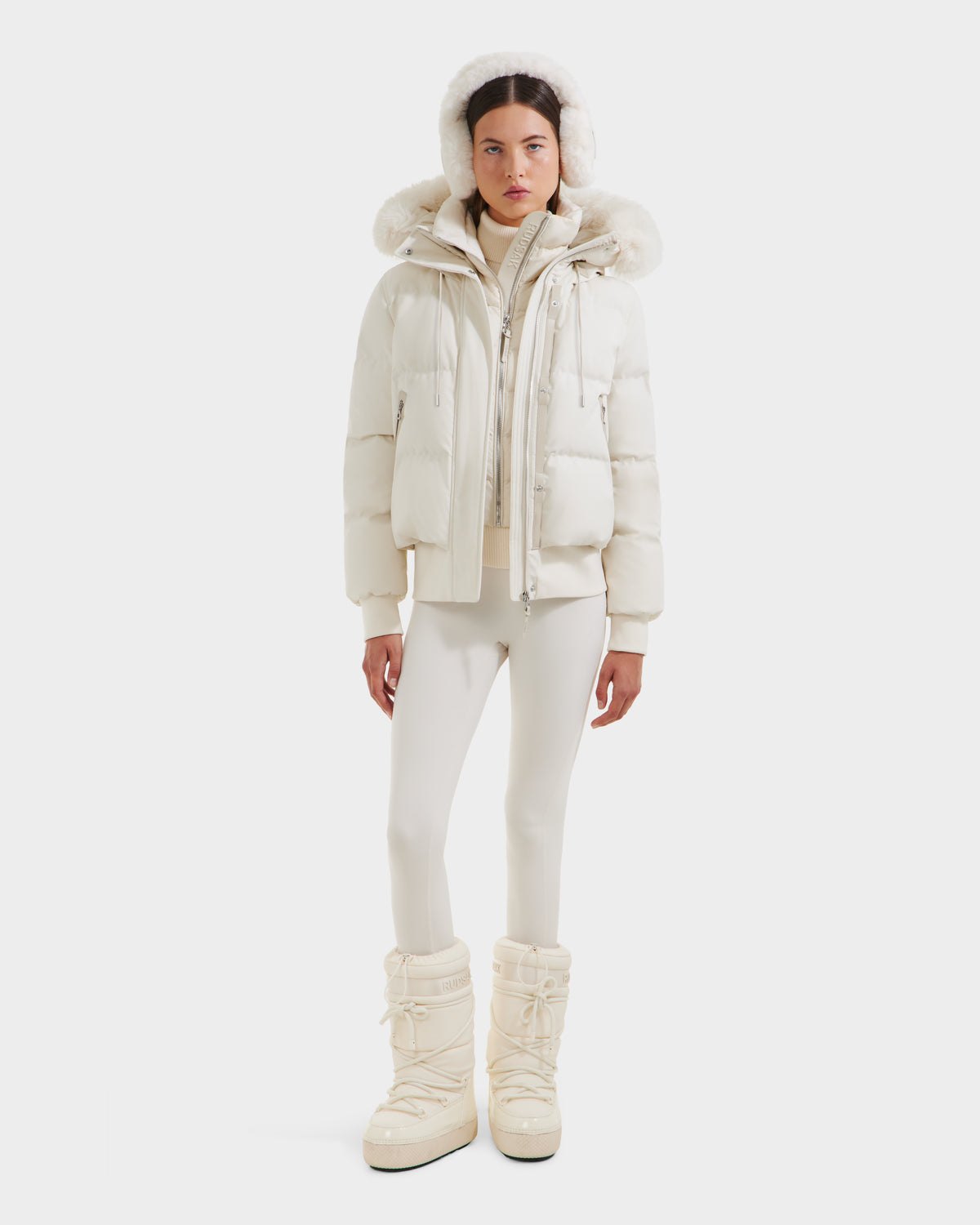 A woman faces forward wearing the Rudsak CIEYNA - 8125579 Vanilla white winter bomber jacket with a fur-lined hood, over a white turtleneck, matching leggings, and tall boots against a plain white background.